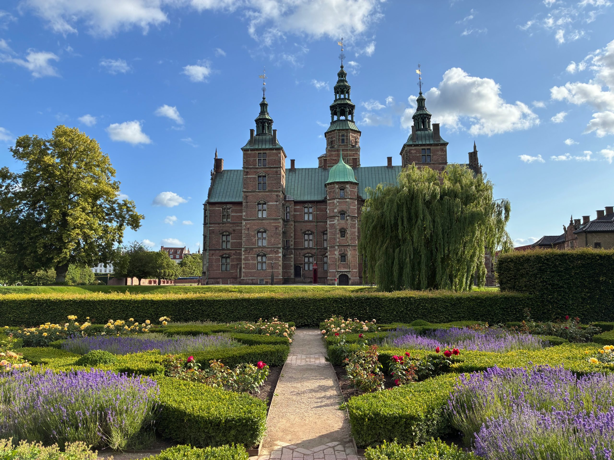 Rosenborg Castle's turrets rise behind manicured garden beds and a willow tree in Copenhagen's King's Garden.