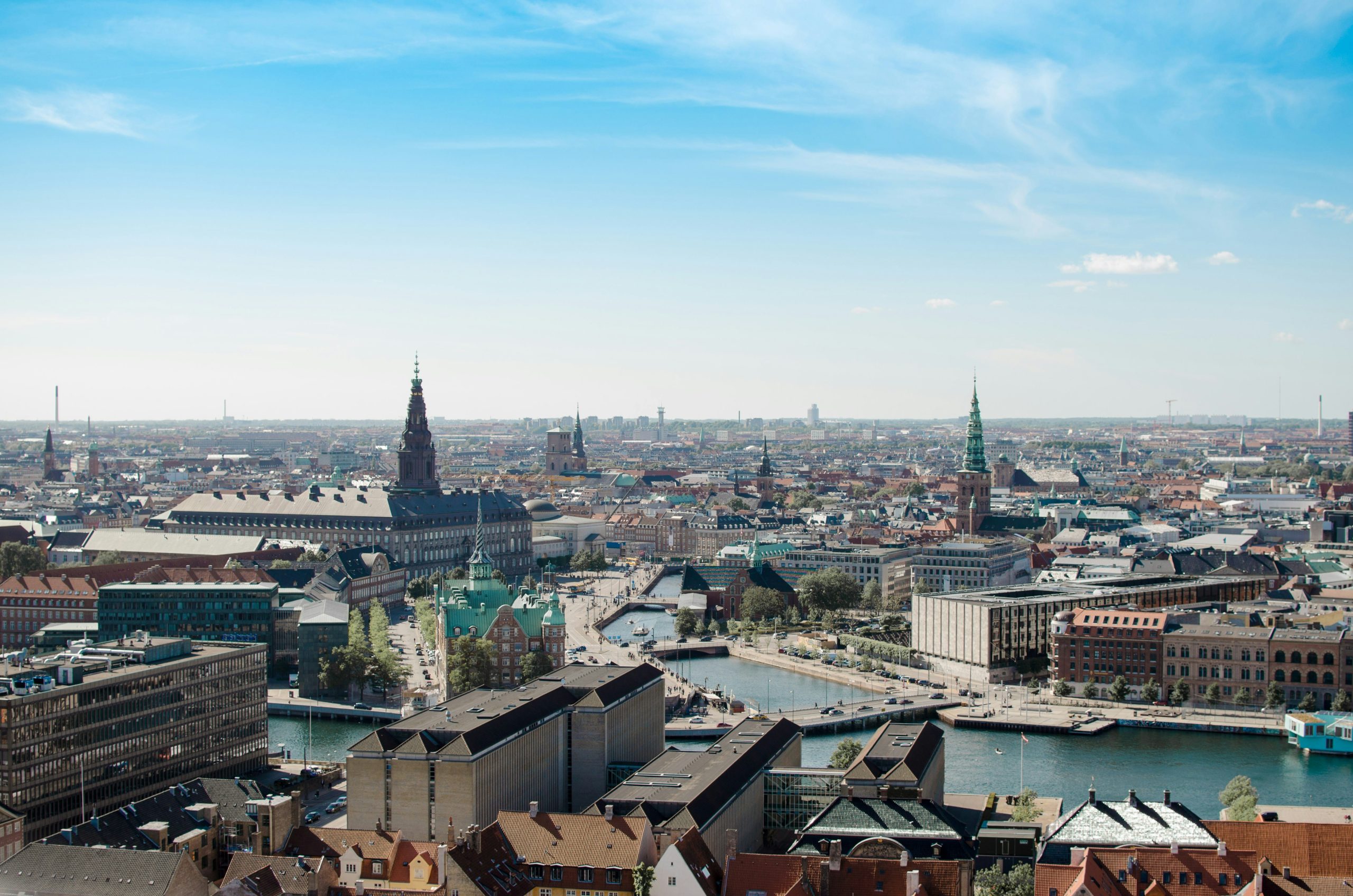An aerial view of central Copenhagen, with a canal cutting through red-roofed buildings and church spires under a blue sky.