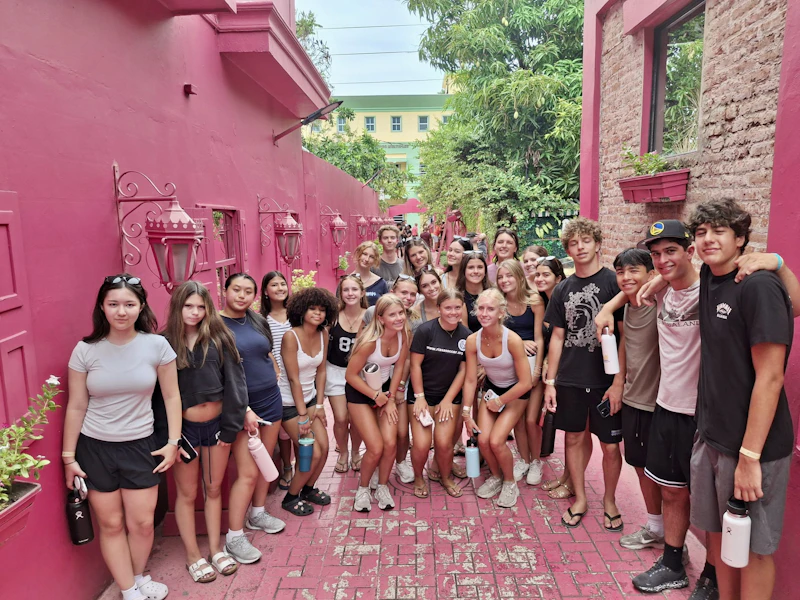Around twenty teen travelers stand along a narrow alley painted entirely bright pink, with lanterns and planters on the walls