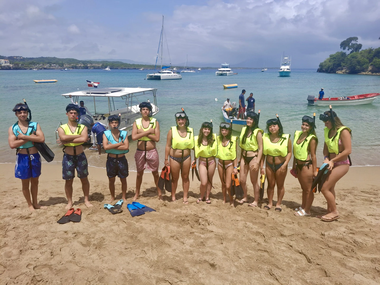 Thirteen teen travelers in yellow buoyancy vests and masks stand on the sand with fins at their feet, boats moored in the bay behind