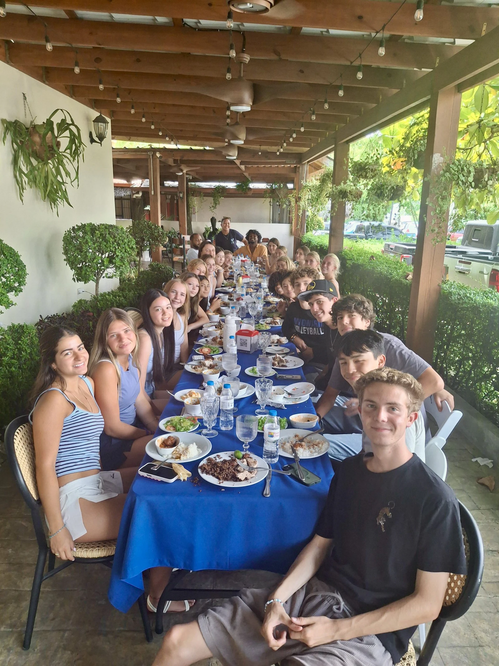 Teen travelers sit along both sides of a long blue-clothed table under a timber pergola strung with lights