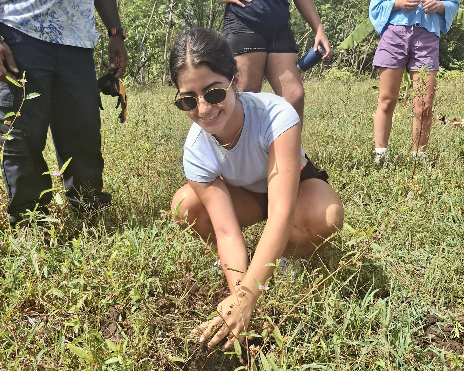 A teen traveler in sunglasses crouches and presses soil around a young plant in rough pasture in the Dominican Republic