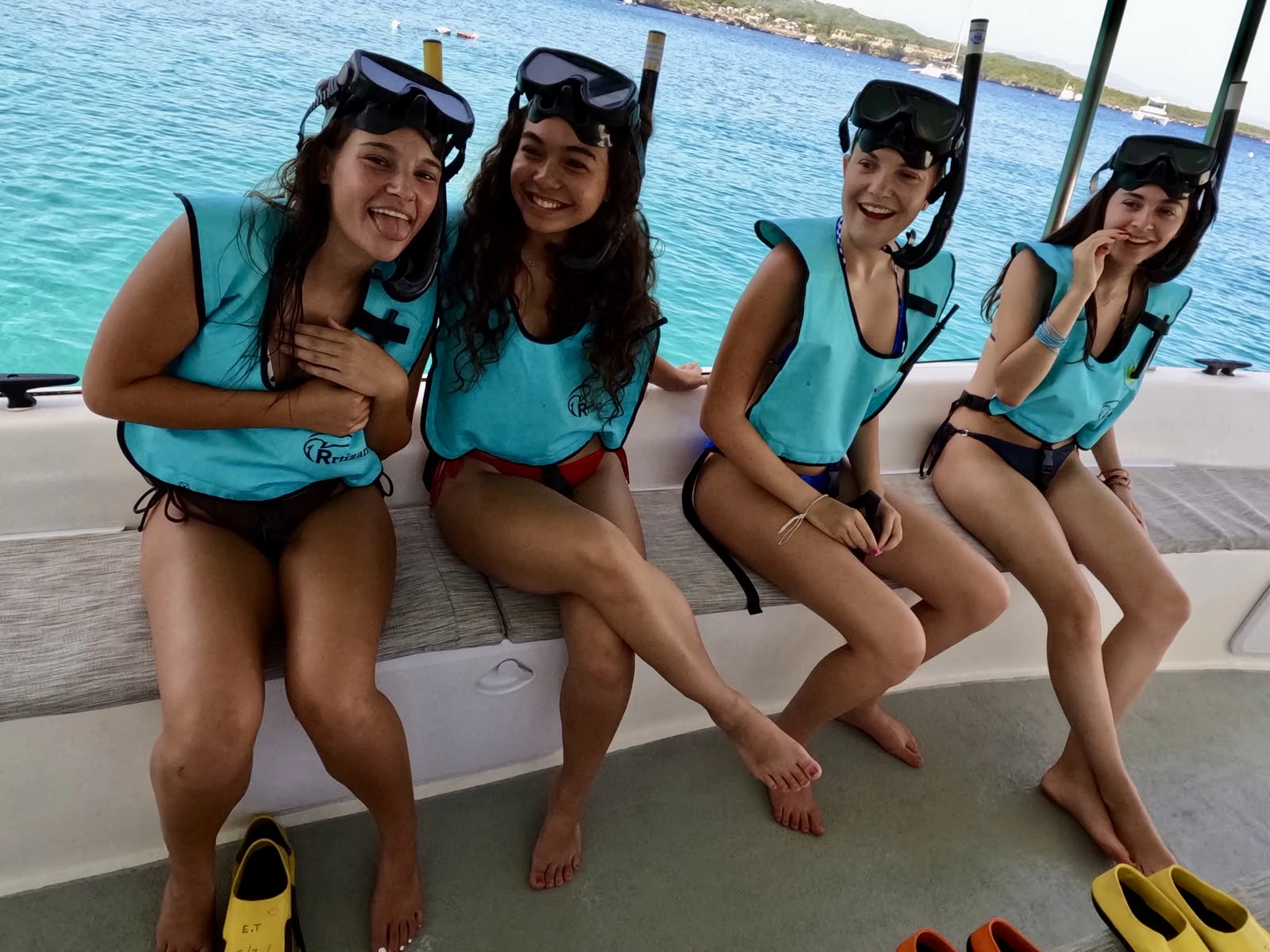 Four teen travelers in turquoise buoyancy vests and snorkel masks sit along the side of a boat on flat blue water