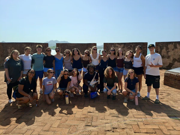Twenty teen travelers stand and crouch on the brick rampart of a colonial fort in the Dominican Republic with the sea behind