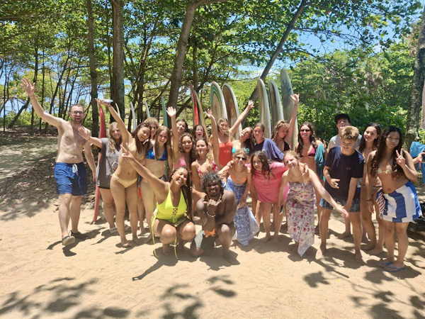 Around twenty teen travelers cheer on the sand in front of a rack of upright surfboards under trees in the Dominican Republic