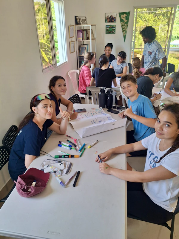Teen travelers sit around white tables with markers and a hand-drawn poster, planning a session in a bright room