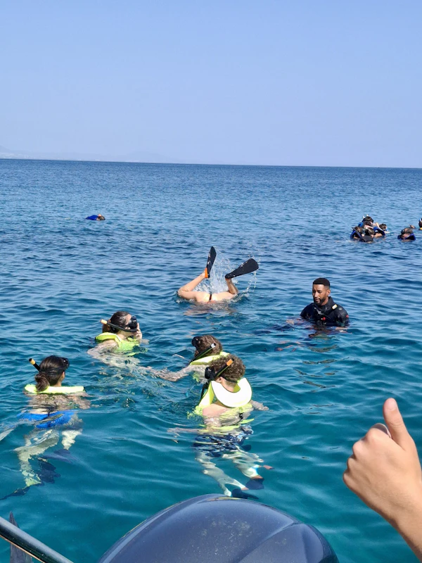 Teen travelers in yellow buoyancy vests float in clear blue water beside a dive boat, one duck-diving with fins in the air