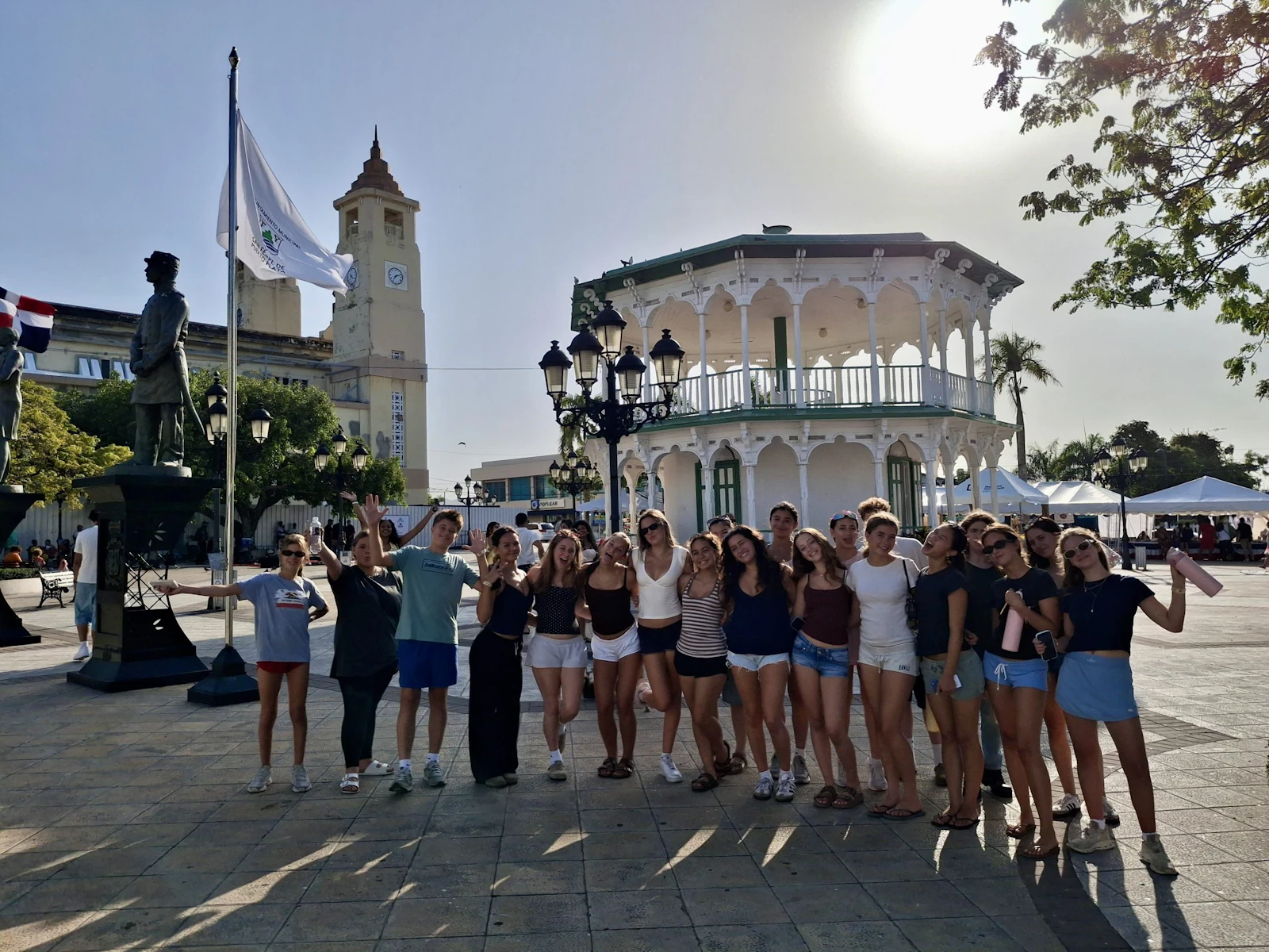 Teen travelers stand in a colonial town square in the Dominican Republic with a white gazebo and a clock tower behind them