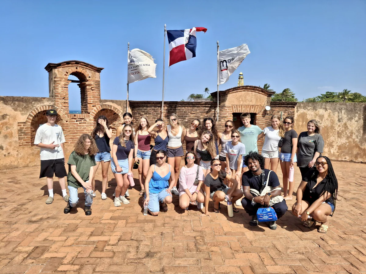 Teen travelers stand in two rows on the brick courtyard of a colonial fort beneath the Dominican flag and two banners