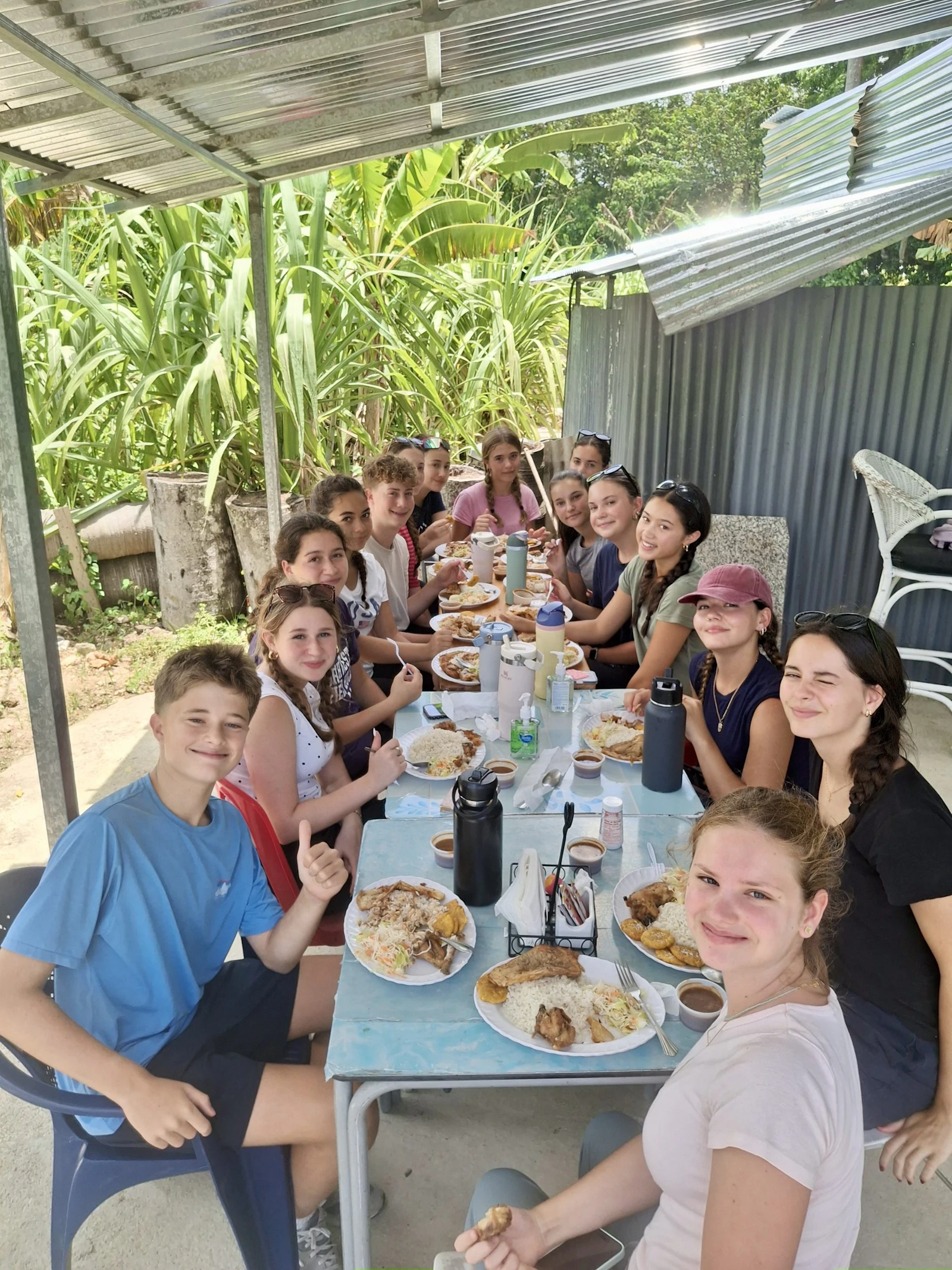 Teen travelers sit around a table under a corrugated shelter eating plates of rice, meat and beans with banana plants behind