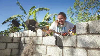 A student smiling as she spreads mortar along the top of a half-built cinderblock wall, palm trees behind her