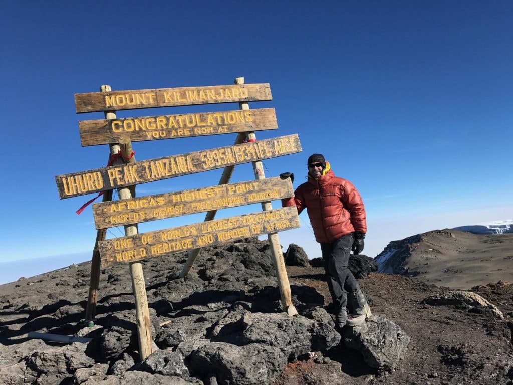 A search and rescue doctor stands beside the summit sign on Mount Kilimanjaro, showcasing the extreme environments where lifesaving missions take place.