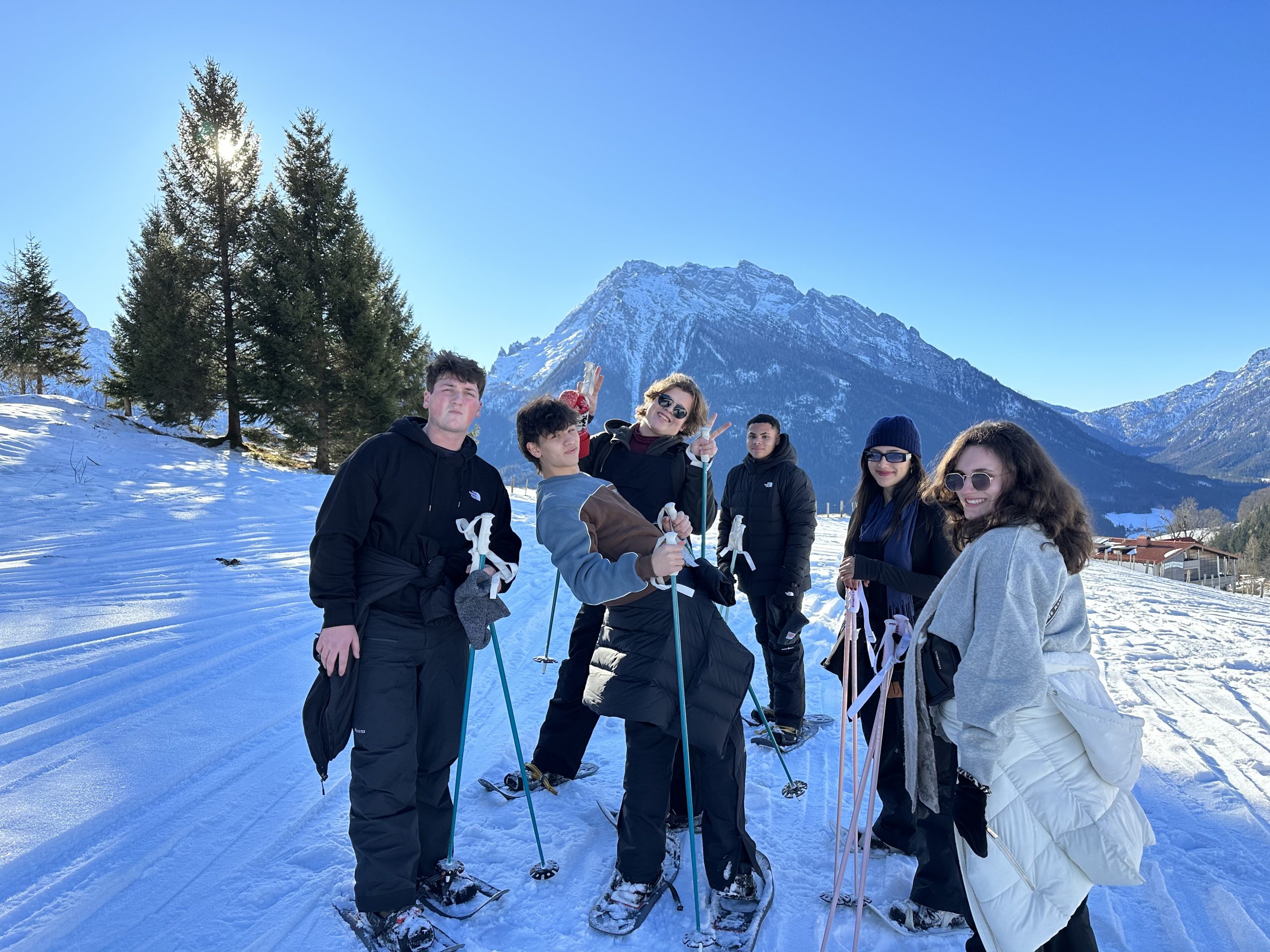 Students with snowshoes and poles pause on a snowy trail beneath the Alps