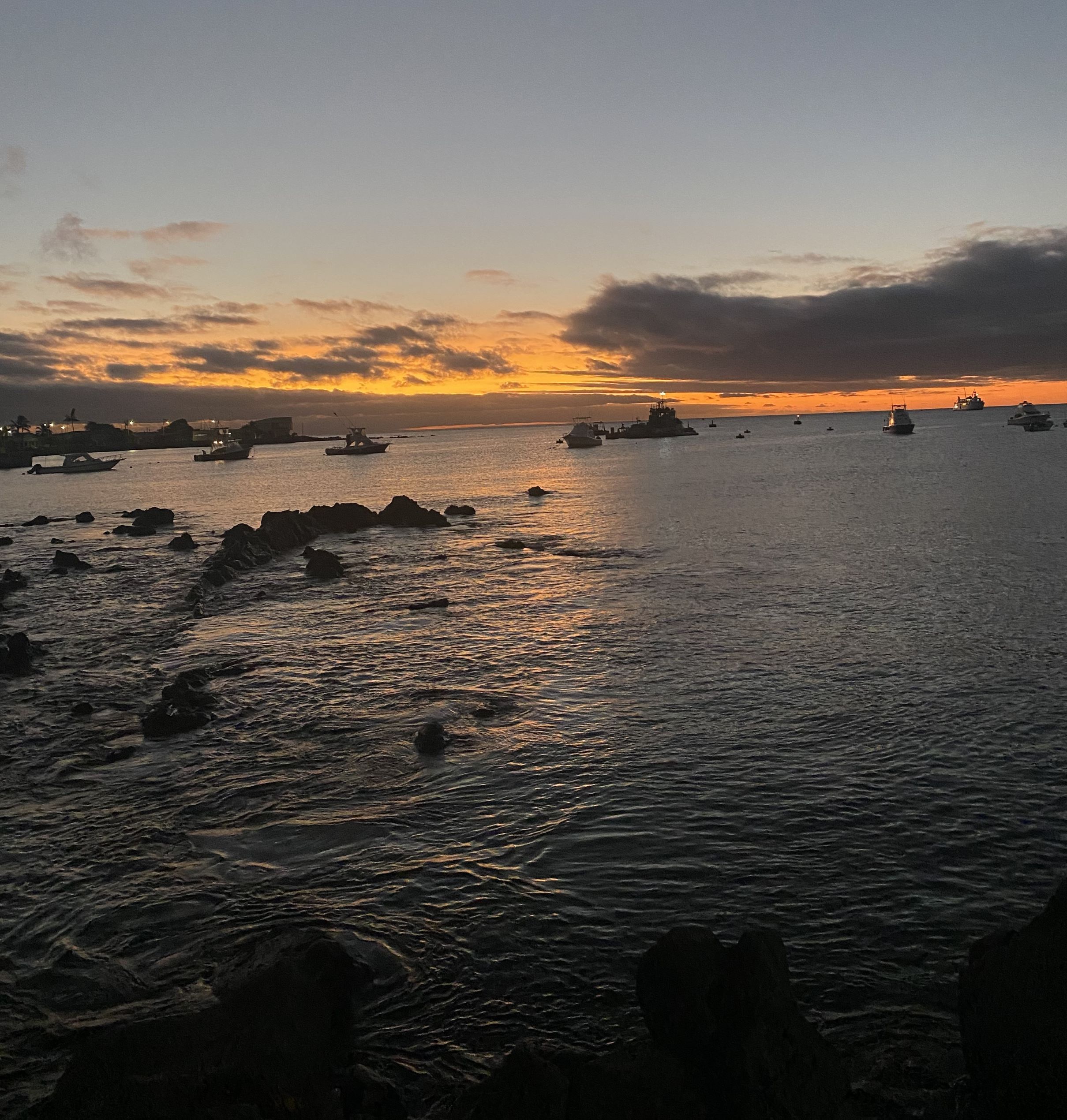 Fishing boats anchor in calm water as the sun sets over rocky shoreline