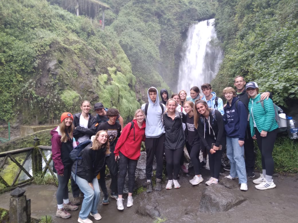 Students pose together near a waterfall surrounded by lush green cliffs