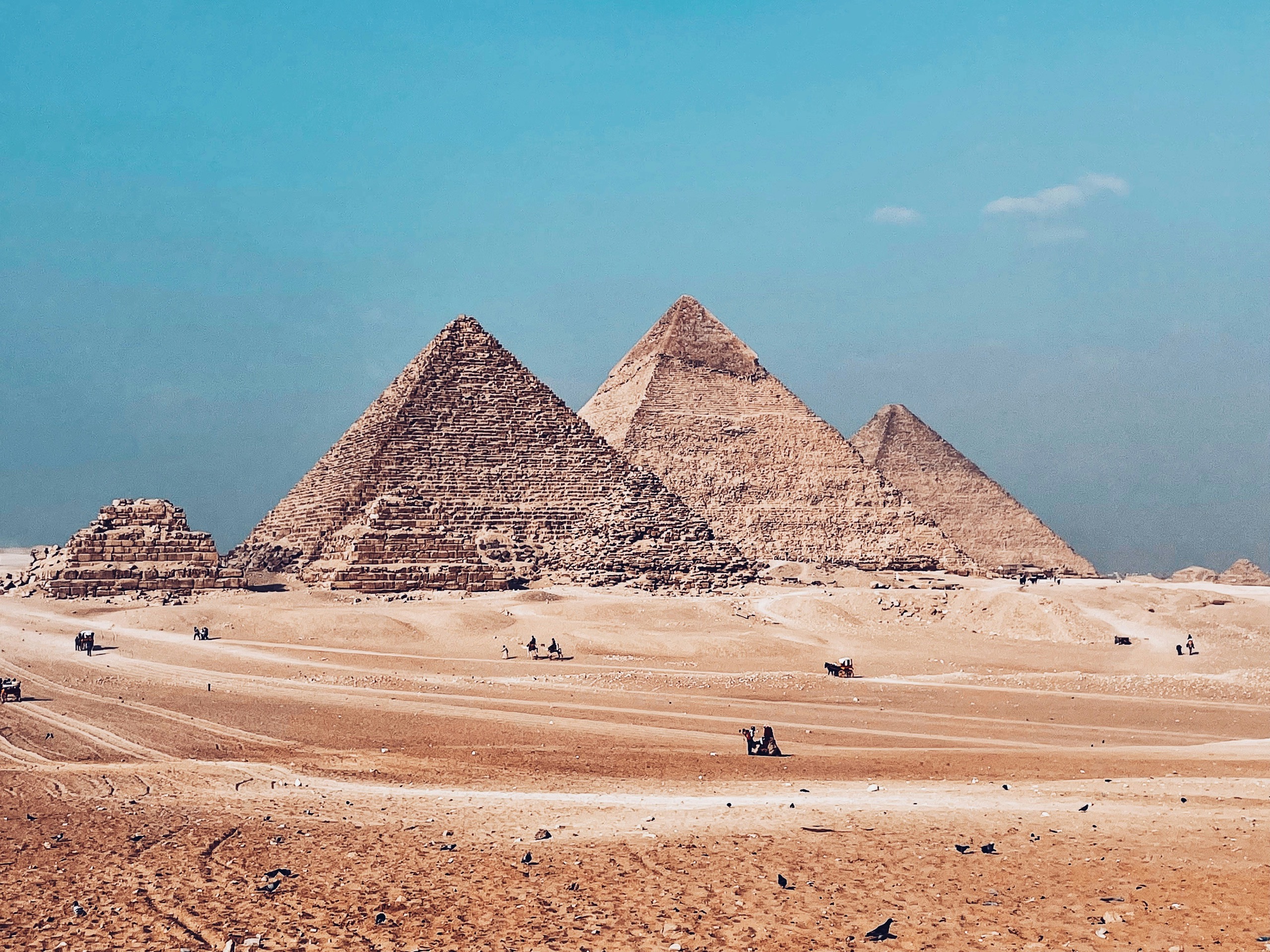 Three pyramids rise from pale desert sand under a clear blue sky, tiny figures crossing the plateau below them.