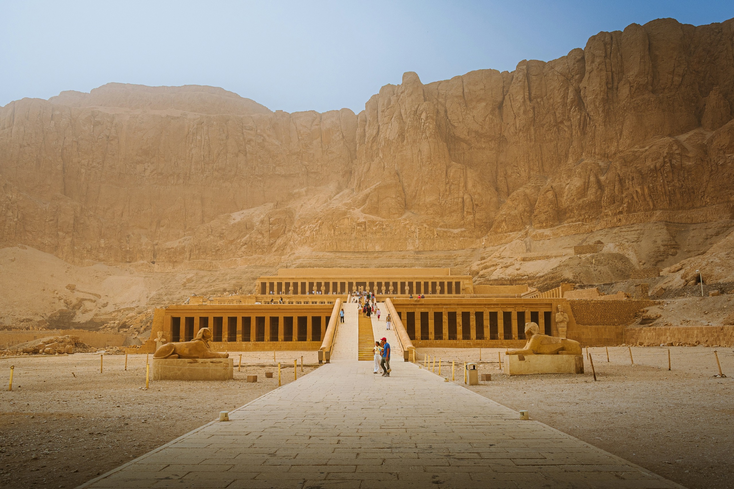 A terraced temple with long colonnades sits at the foot of a sheer desert cliff.