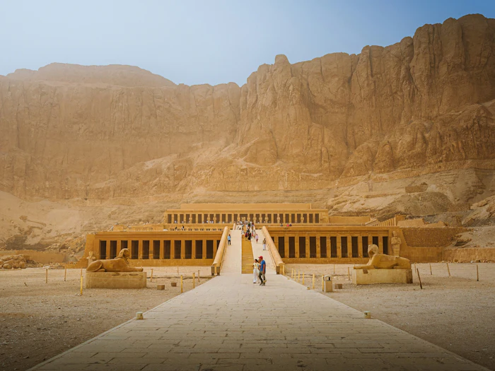 A terraced temple with long colonnades sits at the foot of a sheer desert cliff.