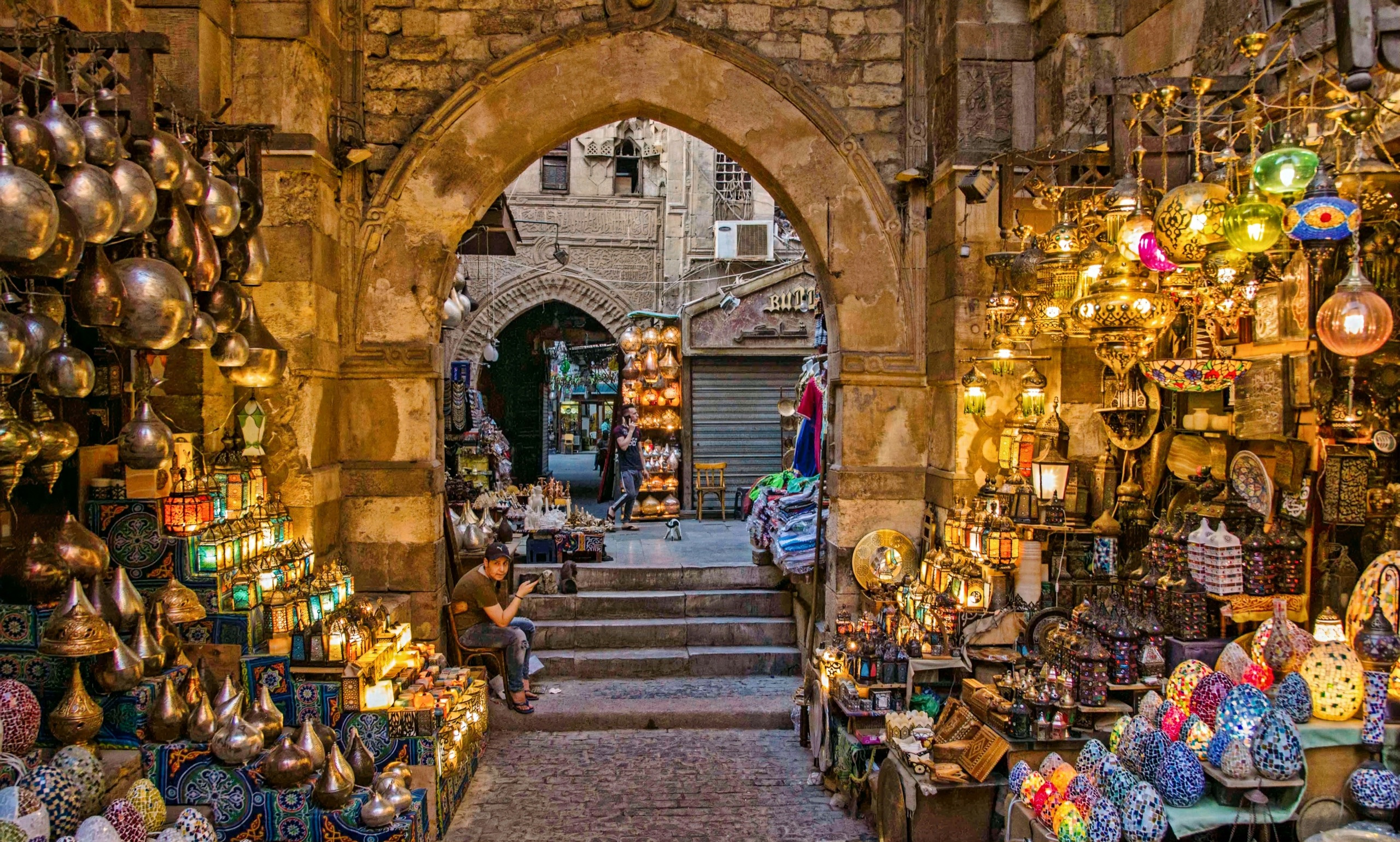 Brass lanterns and painted glass lamps hang in a covered market alley beneath a pointed stone arch.