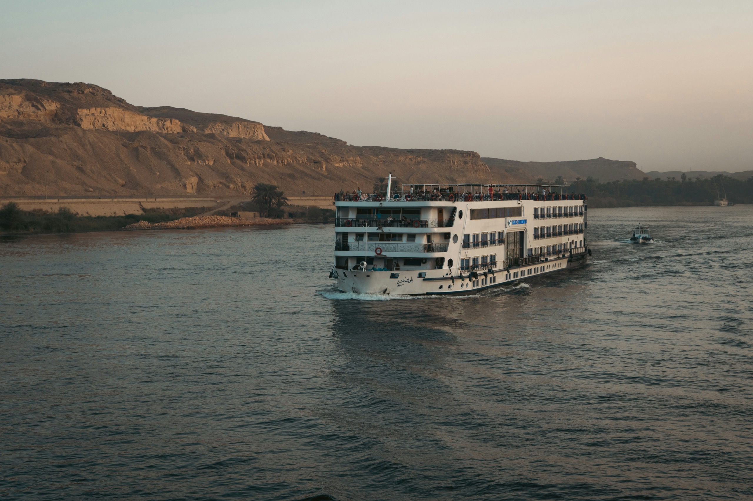 A white river cruise ship sails along a wide river beneath tan desert cliffs.