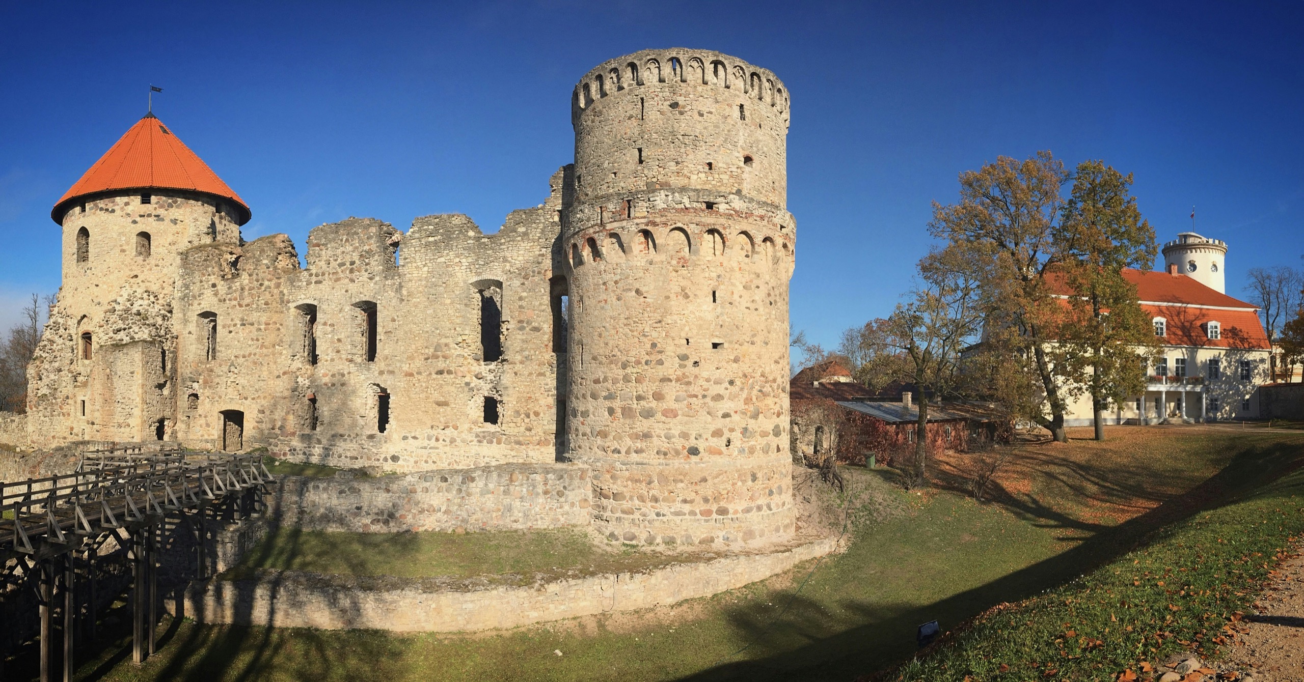 The stone walls and round towers of a ruined medieval castle stand above a lawn, a wooden footbridge crossing the moat.