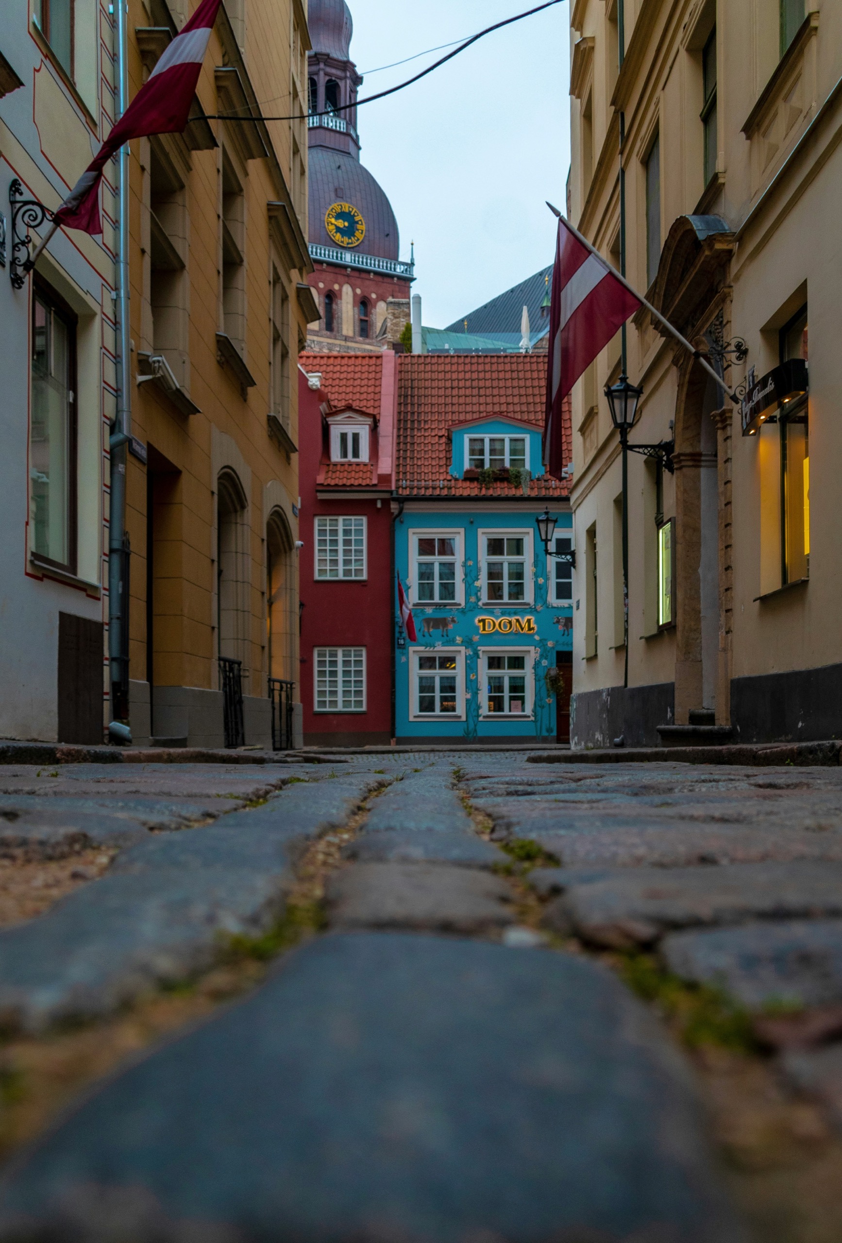A cobbled old-town street between pastel facades, red-and-white flags hanging from the buildings and a clock tower ahead.