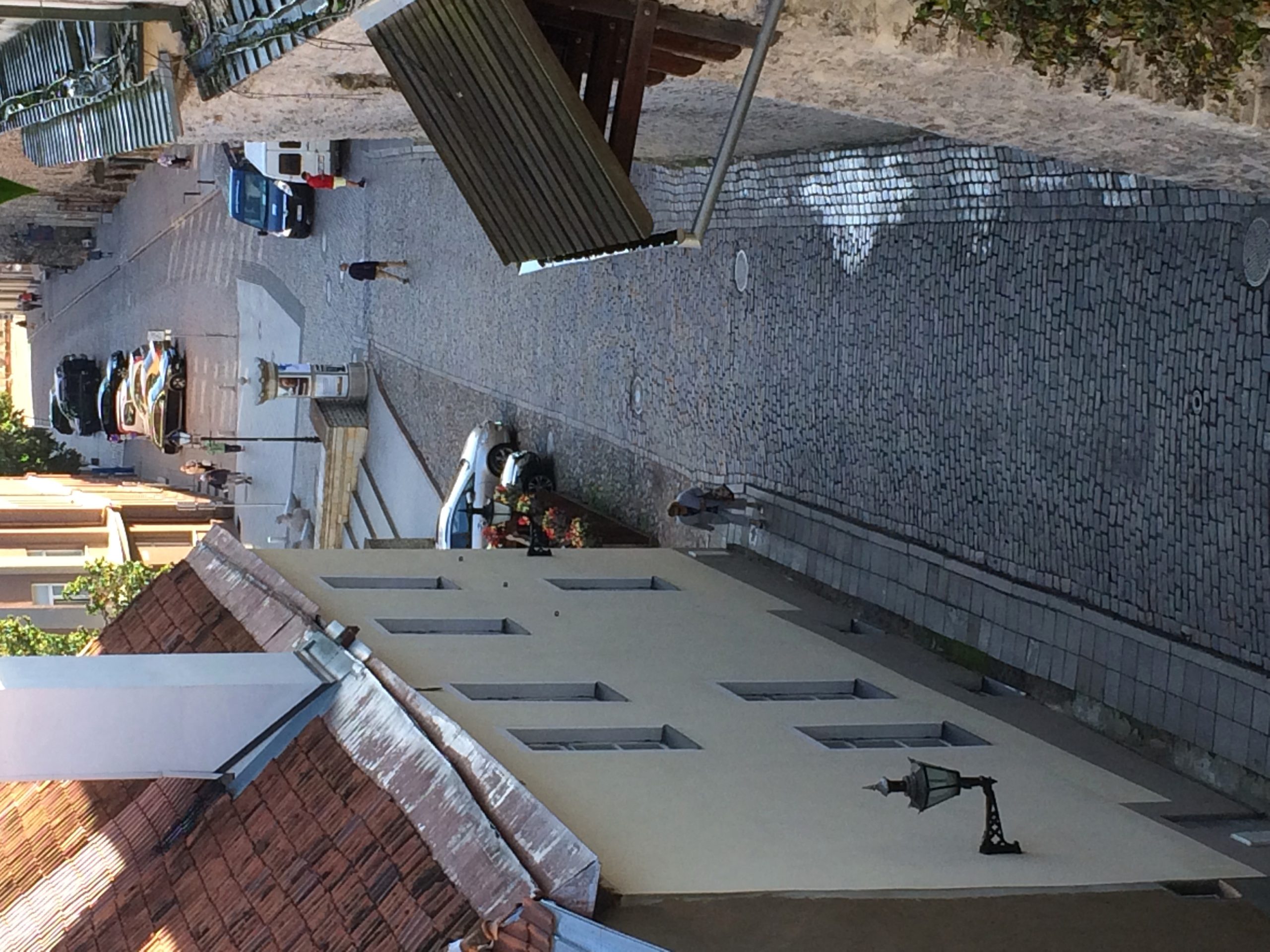 A steep cobbled lane drops past tiled rooftops toward a small square with a few people out walking