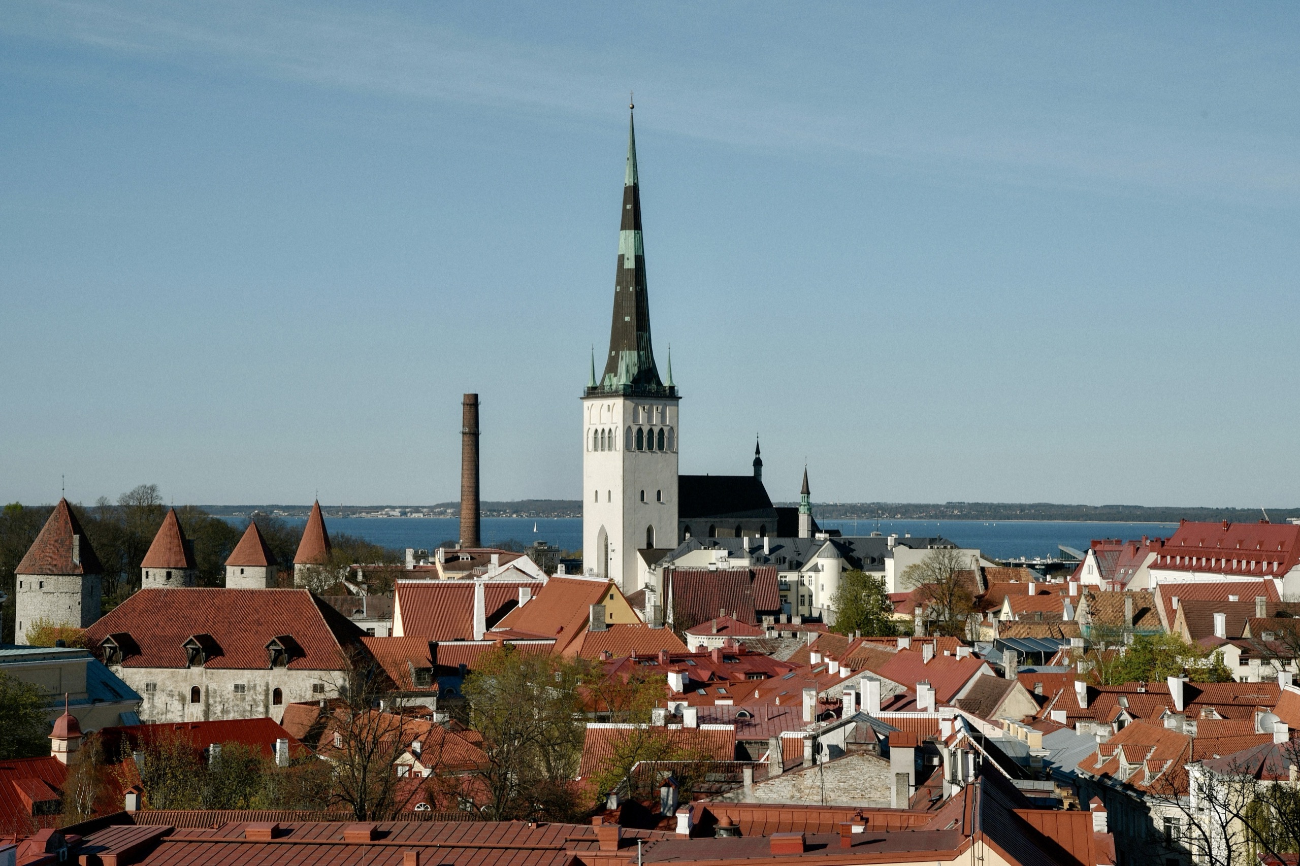 A tall church spire rises above the red-tiled roofs and round stone wall towers of an old town, with the sea beyond.