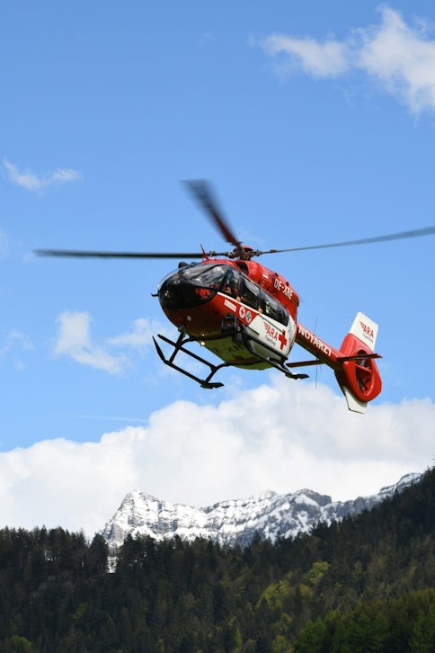 A red helicopter with a first aid symbol painted on the door flying in the mountains with a snowy mountain in the background.