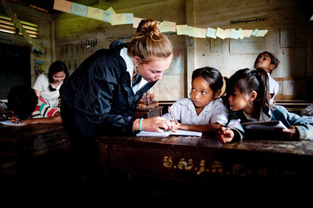 On a school trip, a student helps local schoolchildren with writing at a classroom desk