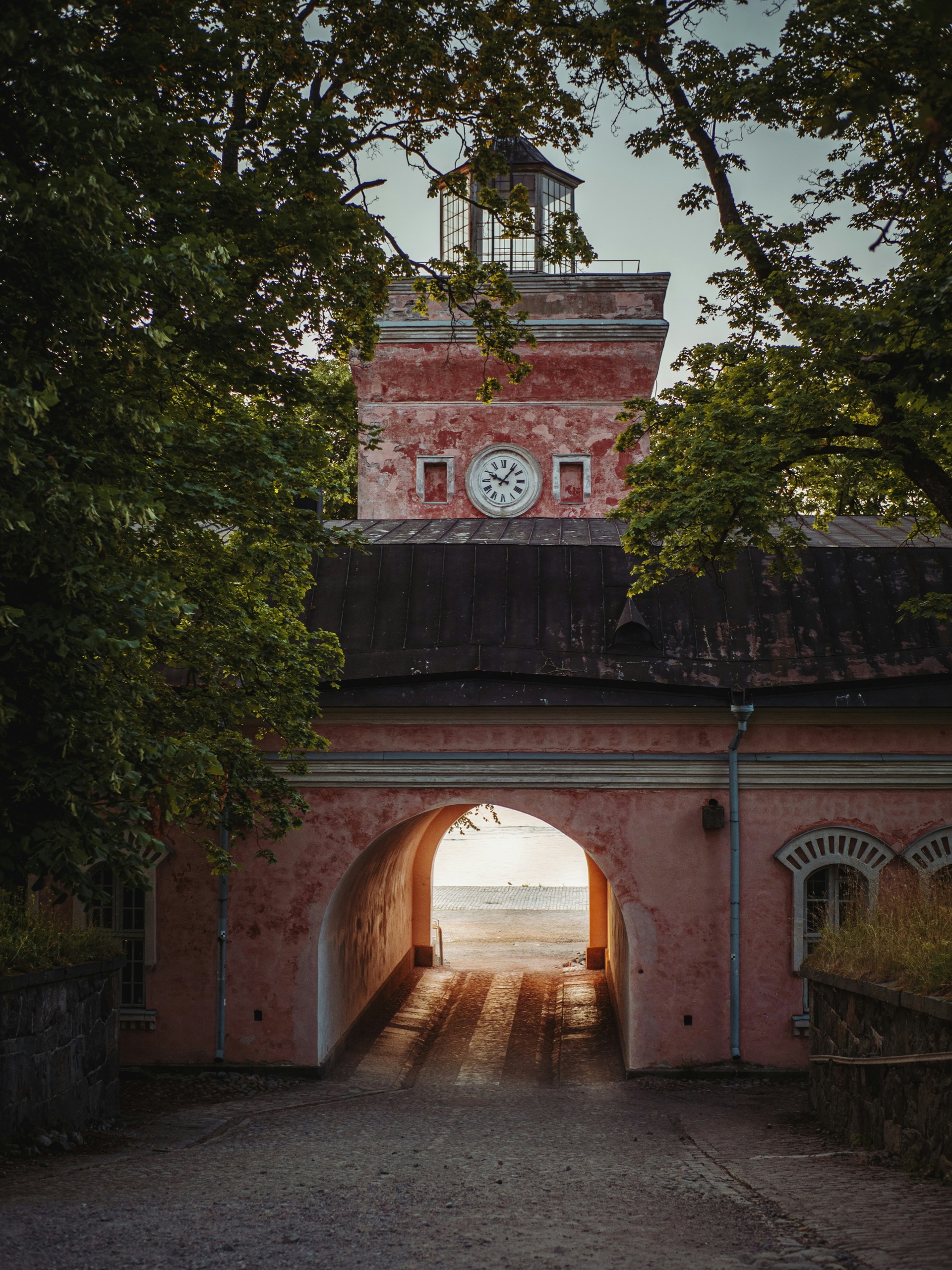 The pink gatehouse of Suomenlinna's fortress, a clock face under its lantern tower, seen through an arched passage framed by summer trees.