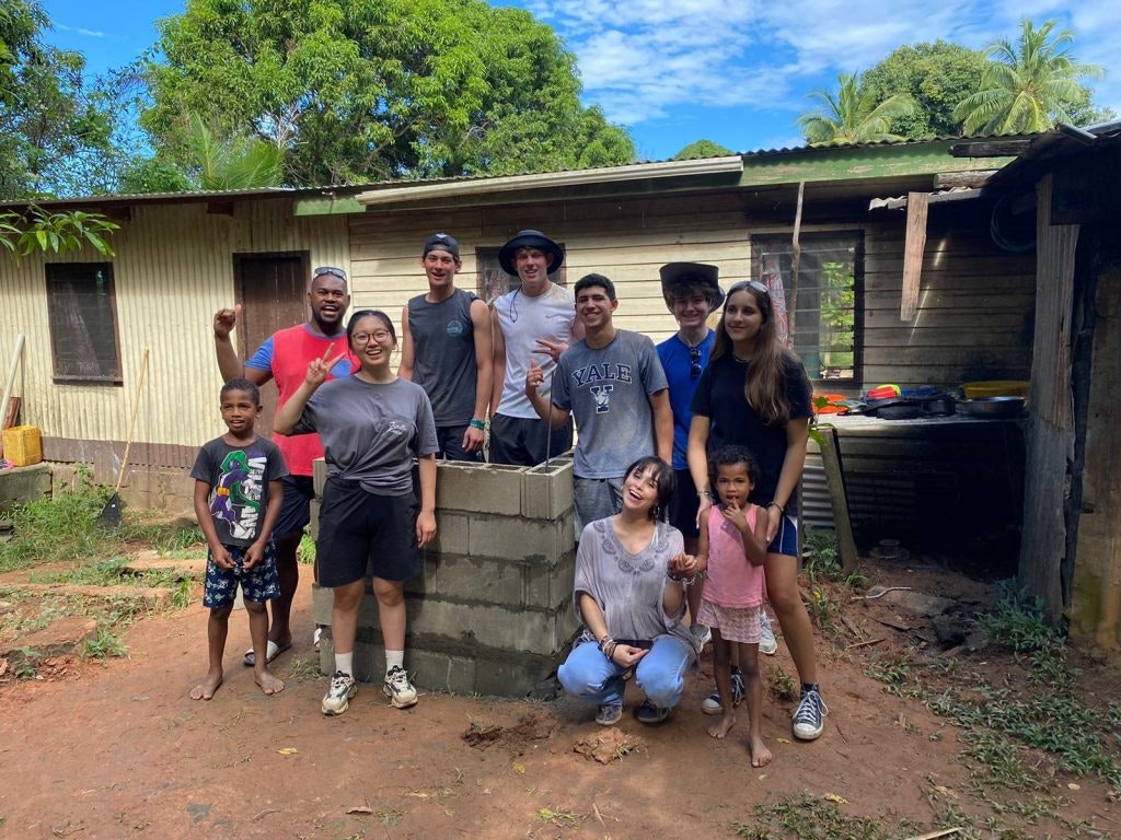 In this photo, a diverse group of cheerful volunteers poses proudly in front of a rustic wooden house, celebrating their successful community service project as they stand around a newly constructed brick structure under a bright blue sky.