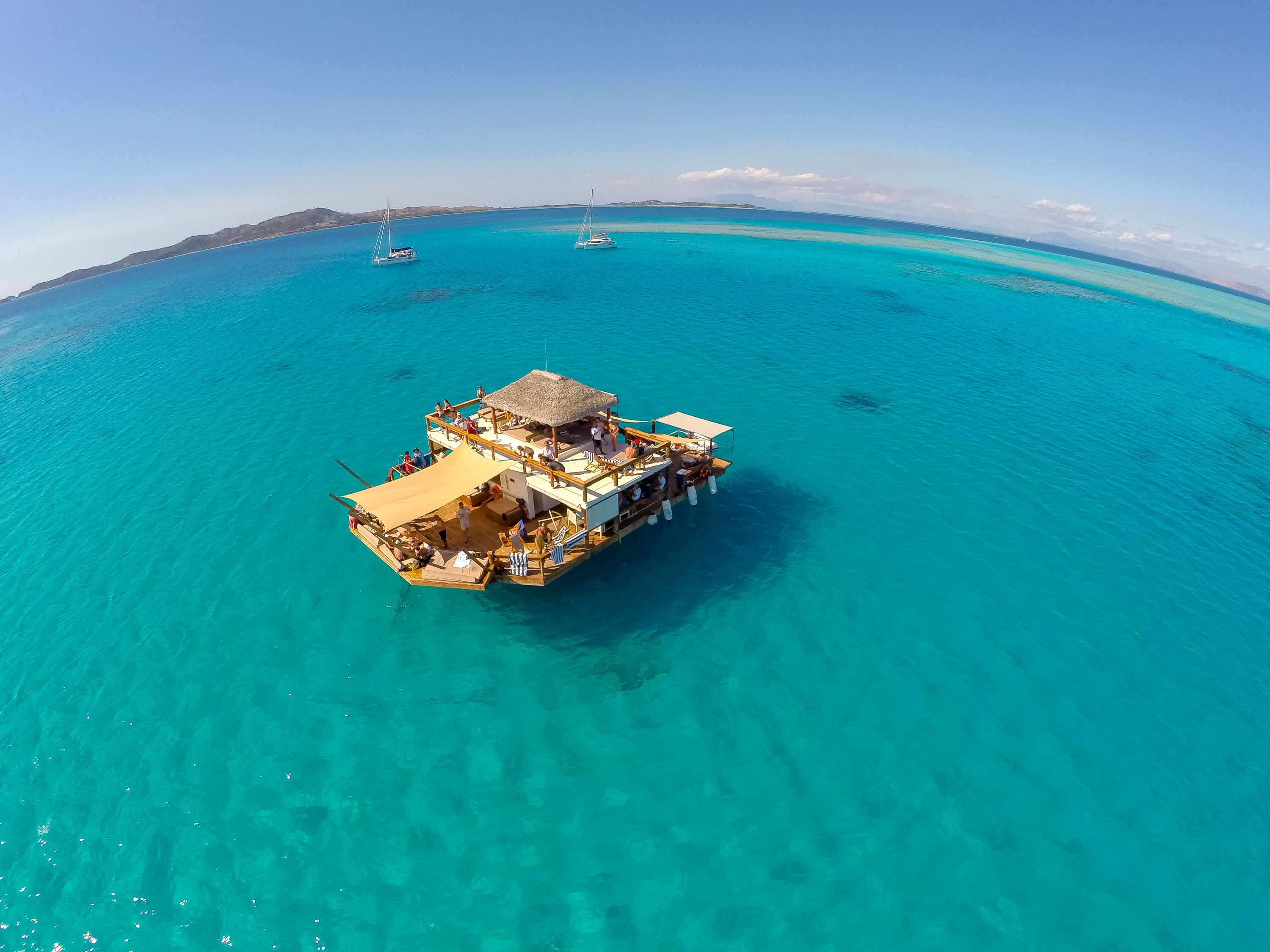 An overwater platform bar sits amid turquoise lagoon waters with sailboats in the distance