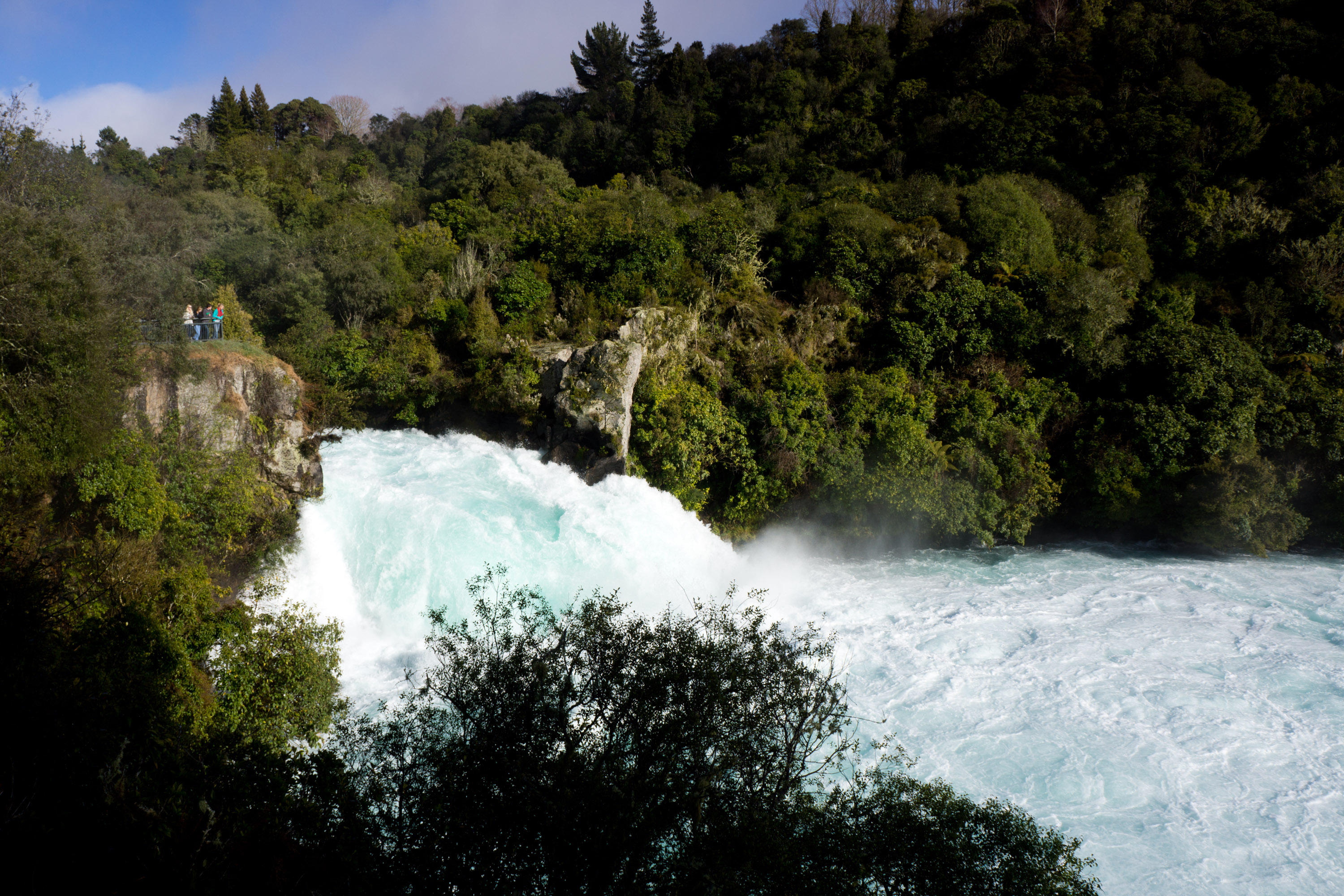 A powerful waterfall rushes through a forested gorge as onlookers watch from a cliffside viewpoint