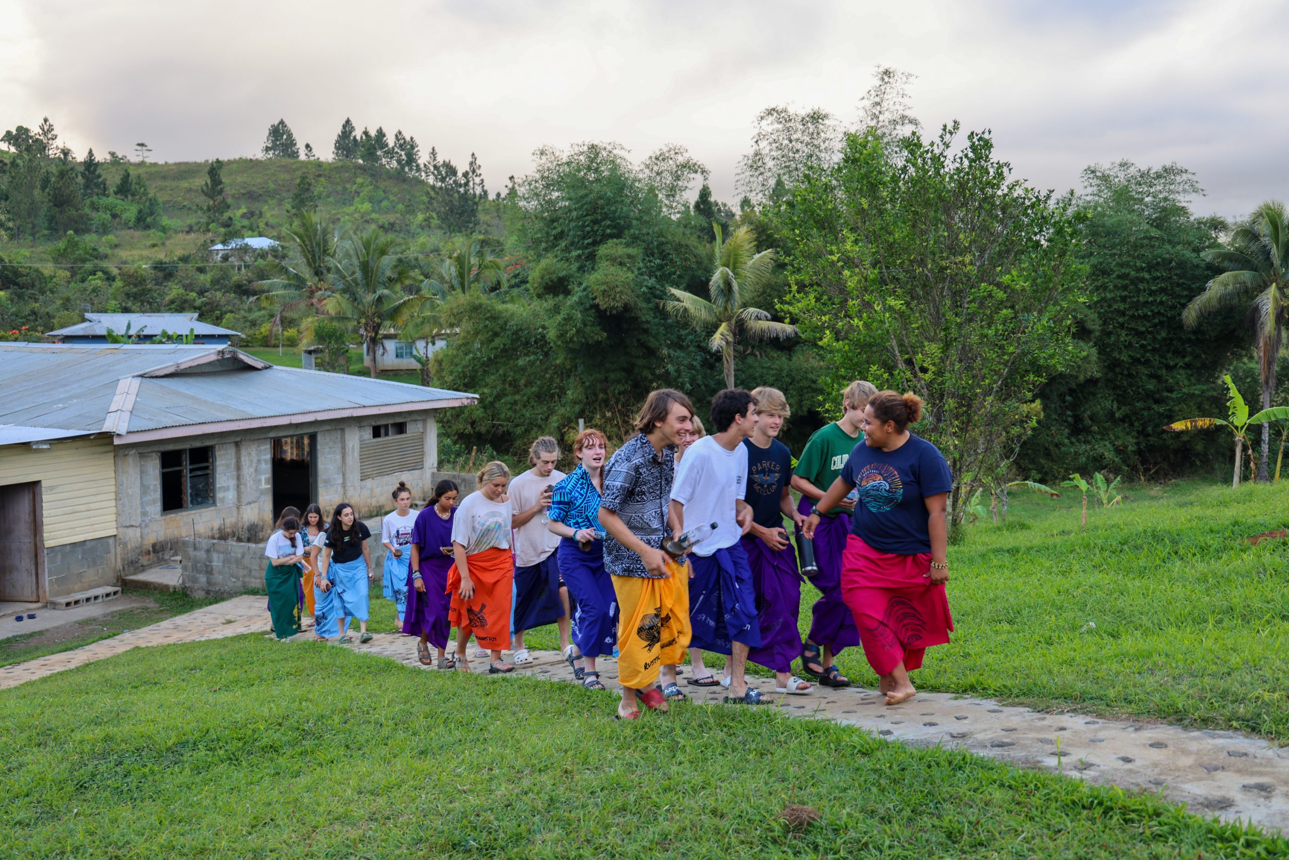Students in traditional wrap skirts walk together along a path near village buildings