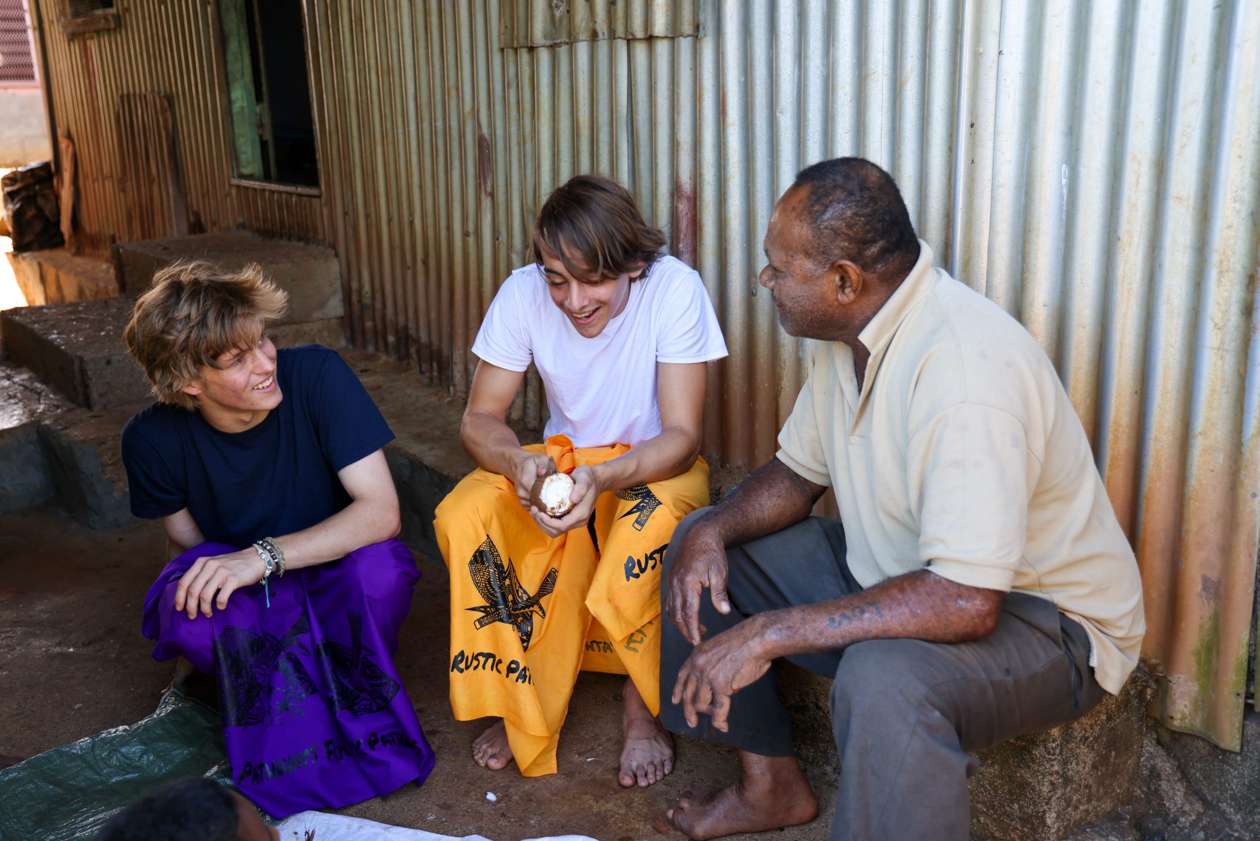 Students sit and talk with a local host outside a corrugated metal building in Fiji