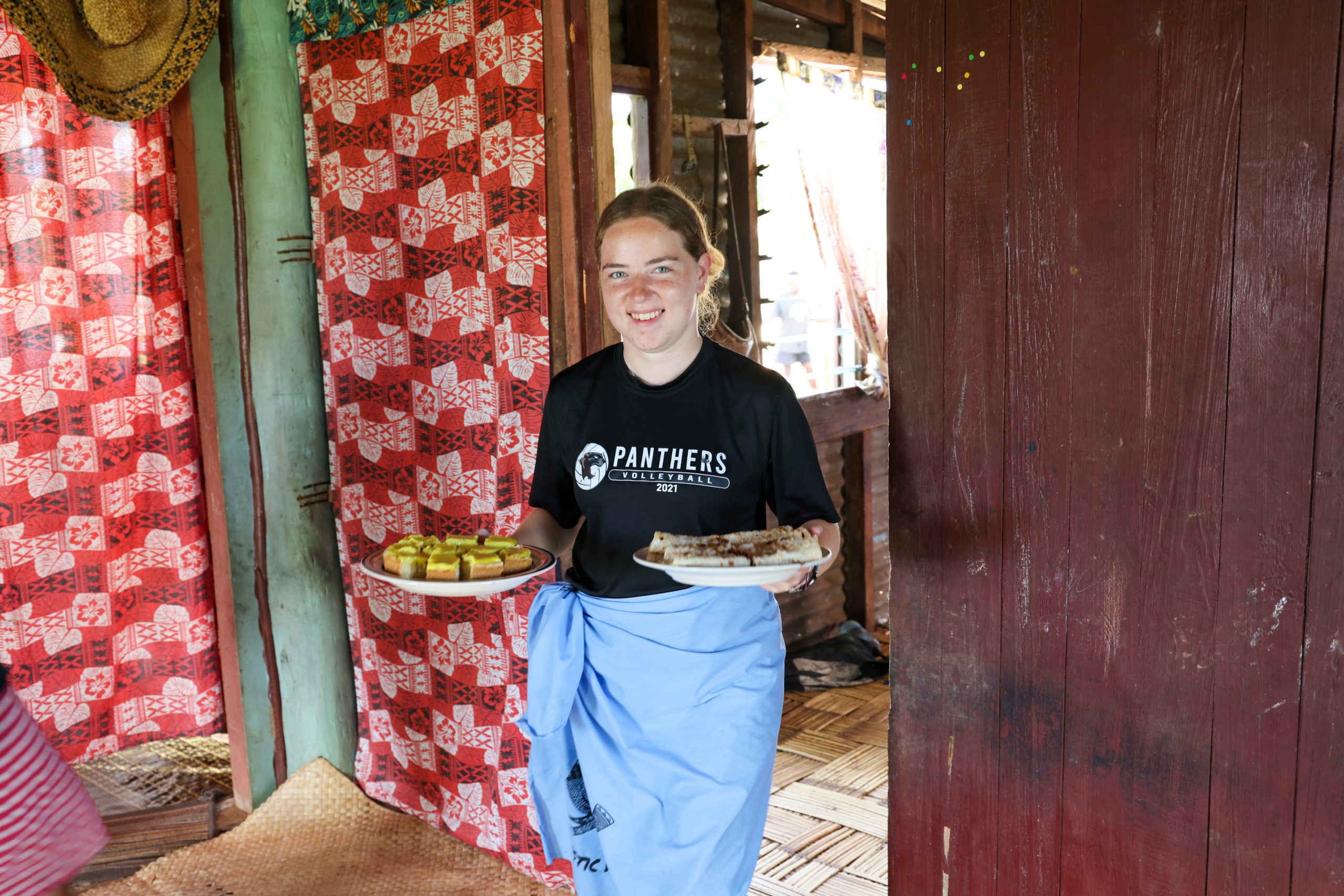 A student carries plates of food through a wooden building decorated with red patterned fabric