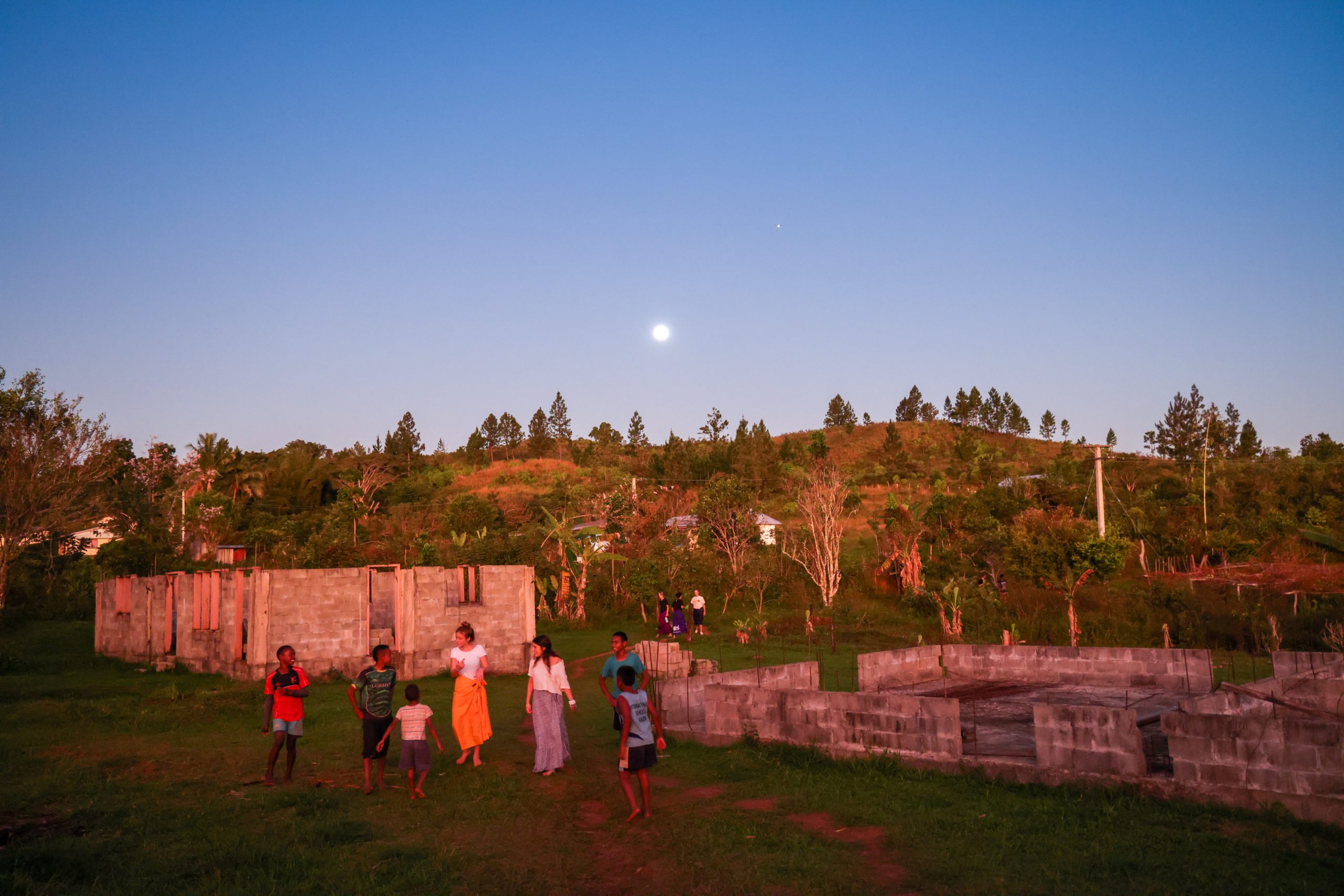 Students and villagers gather in a grassy yard at dusk near stone walls with the moon rising over the hills