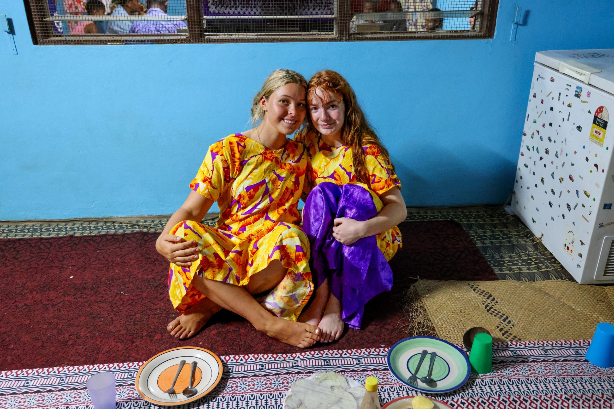 Students in matching floral Fijian dresses sit together on a woven mat indoors