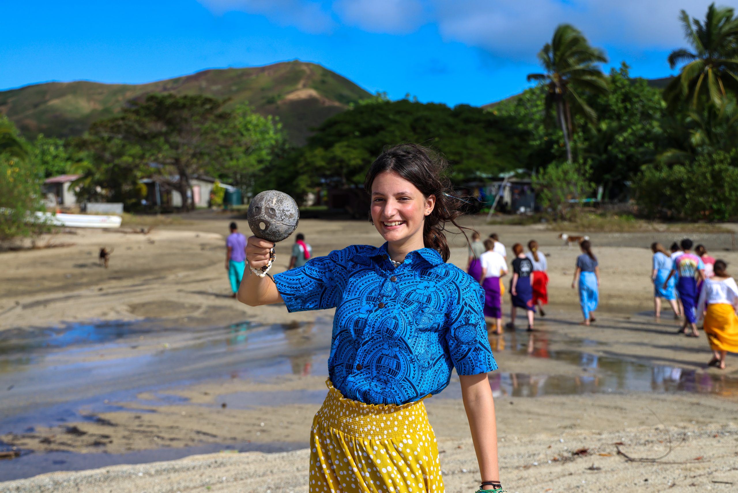A student holds up a shell on a Fijian beach with hills and palm trees behind