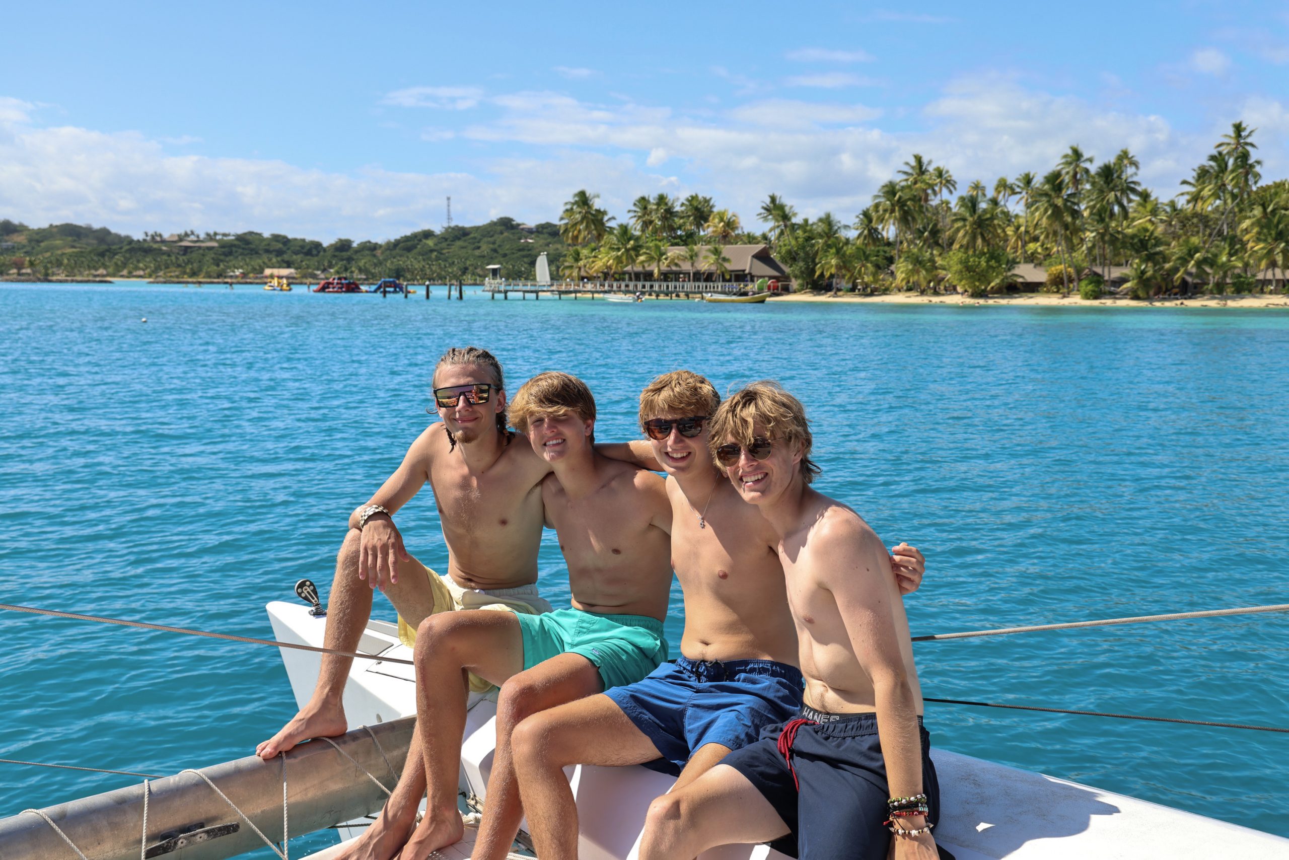 Students sit on a boat's bow with a palm-lined island shore behind them