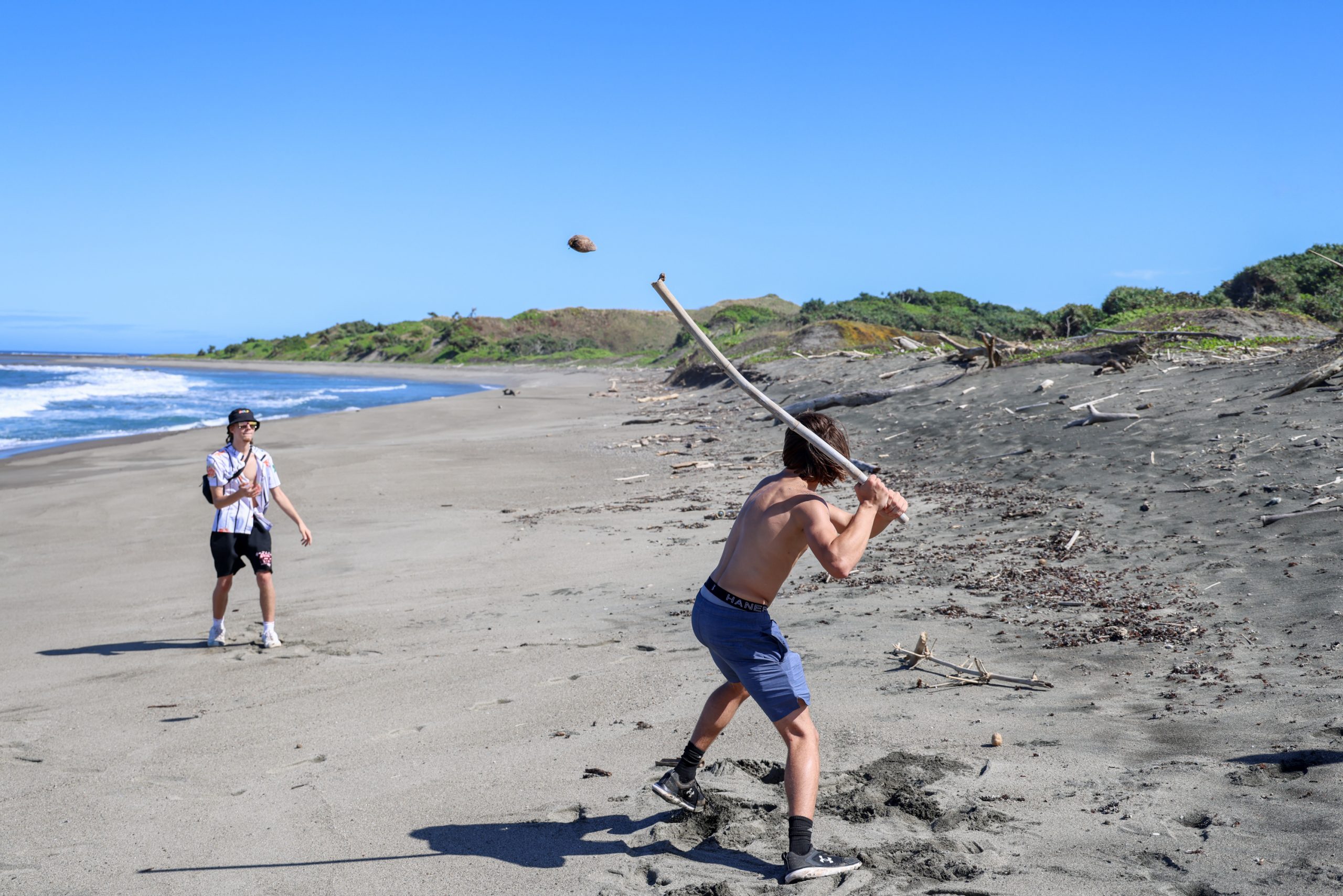 A student swings a driftwood stick at a coconut on a beach