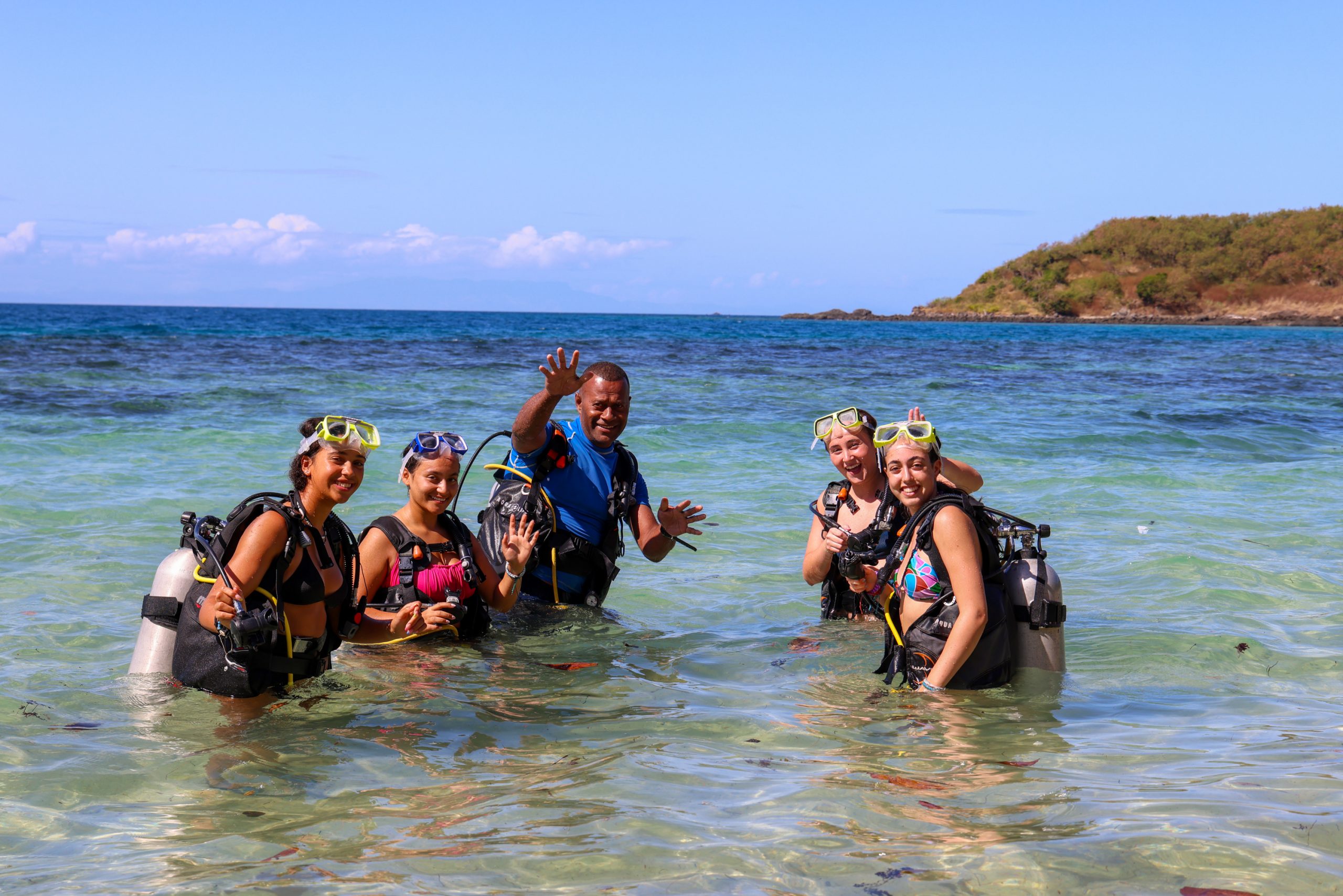Students in scuba gear wave from shallow water with an island in the background