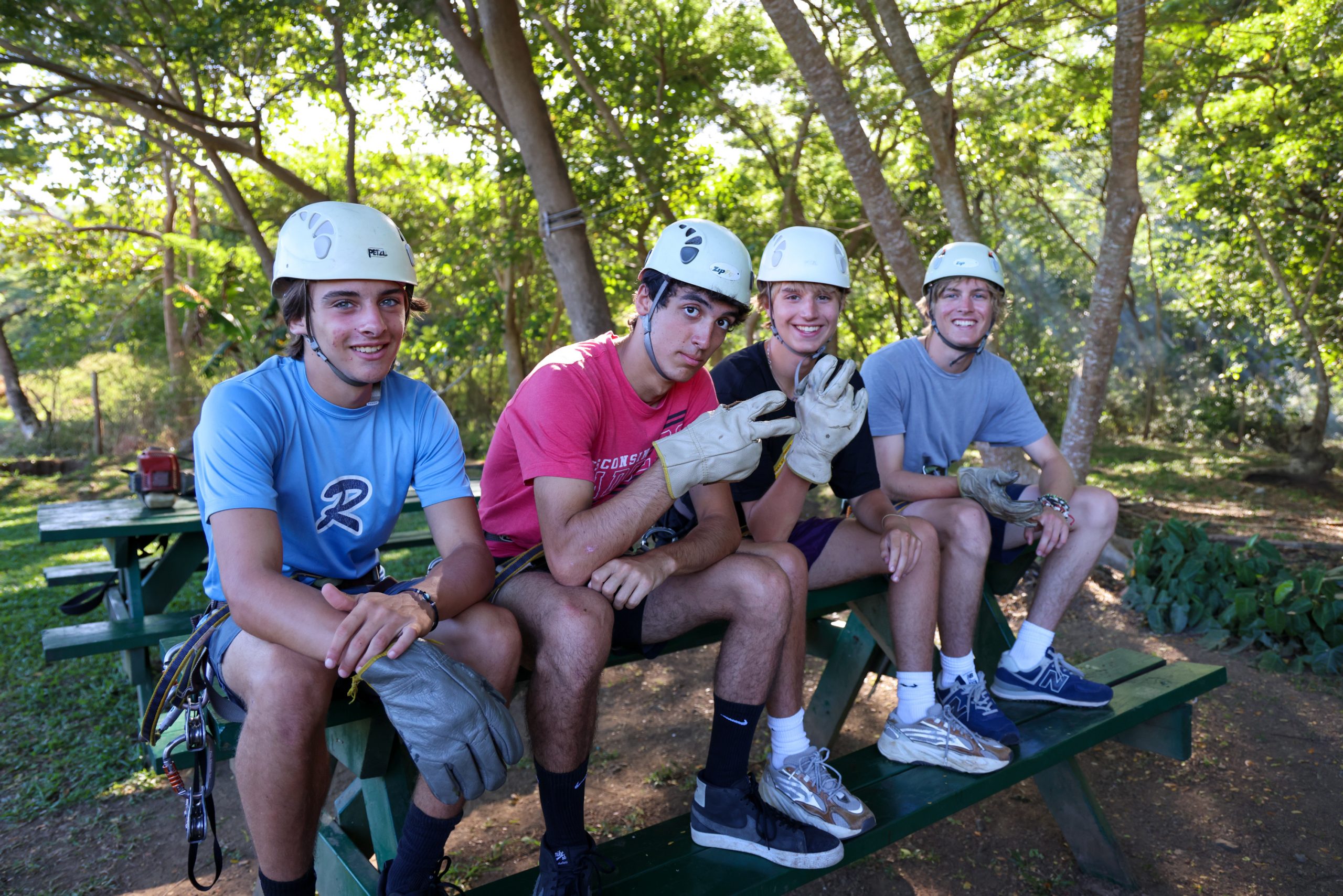 Students in zipline helmets sit together on a picnic table in the forest