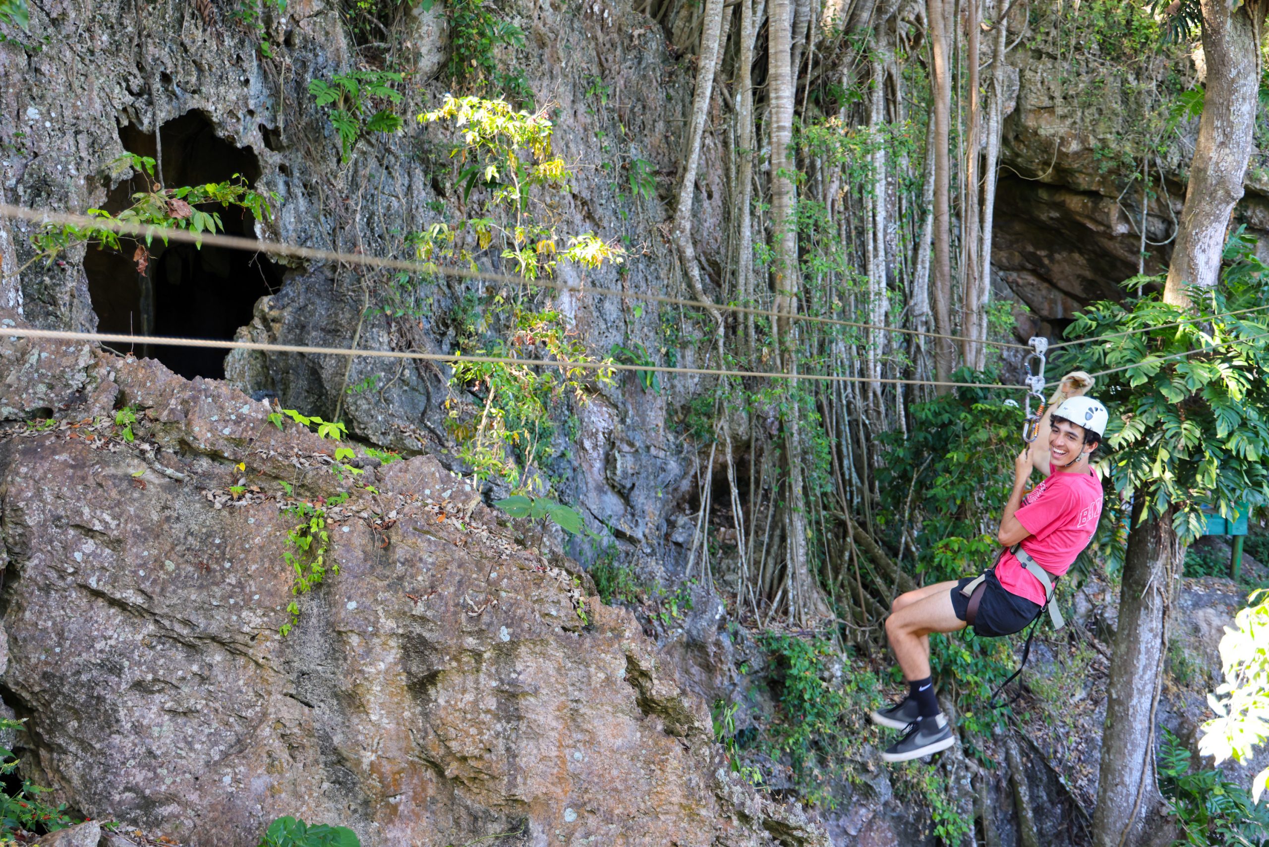 A student ziplines past a rocky cliff face draped in vines
