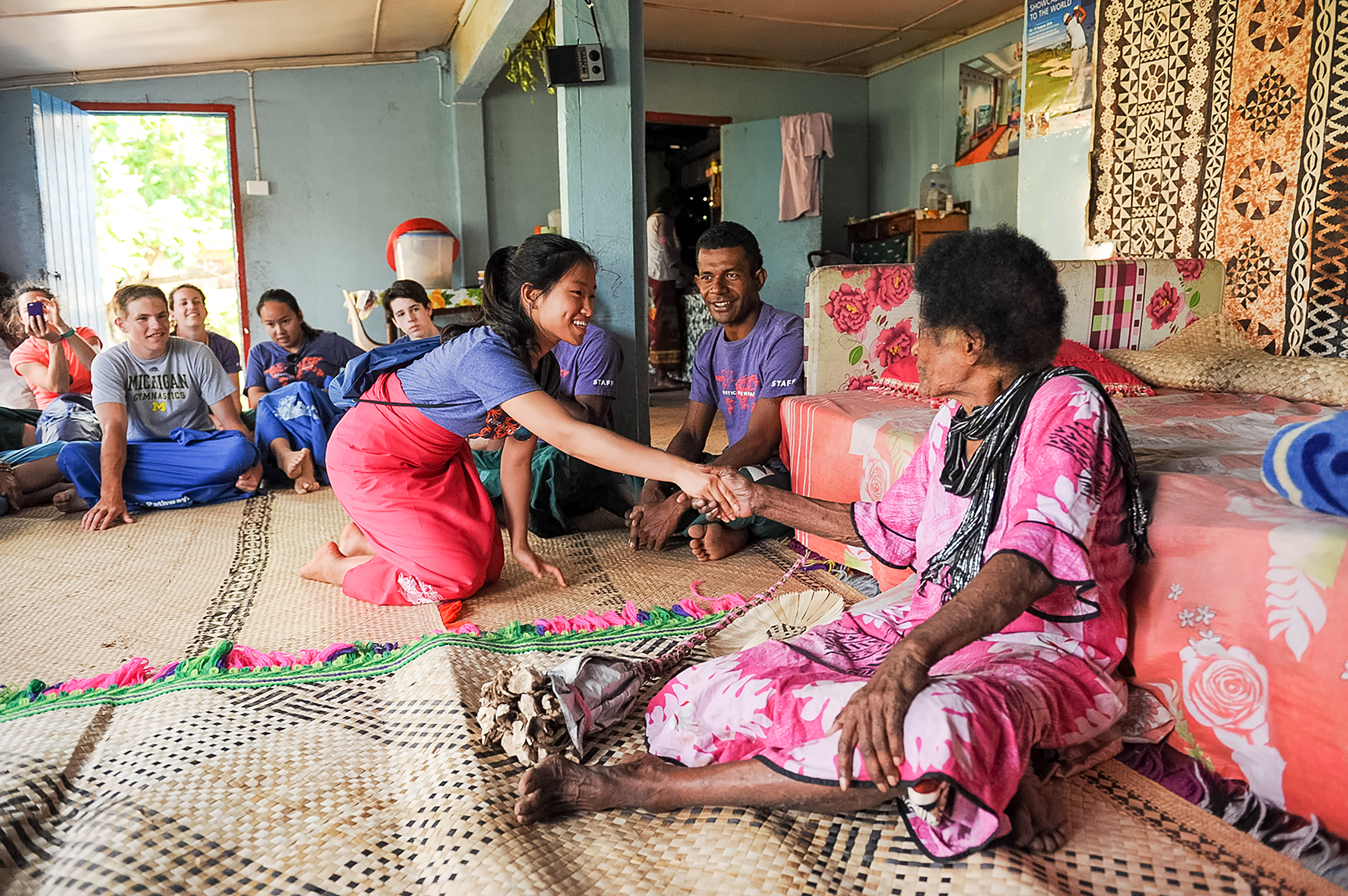 Student travelers greet an elder while seated on a woven mat inside a Fijian home