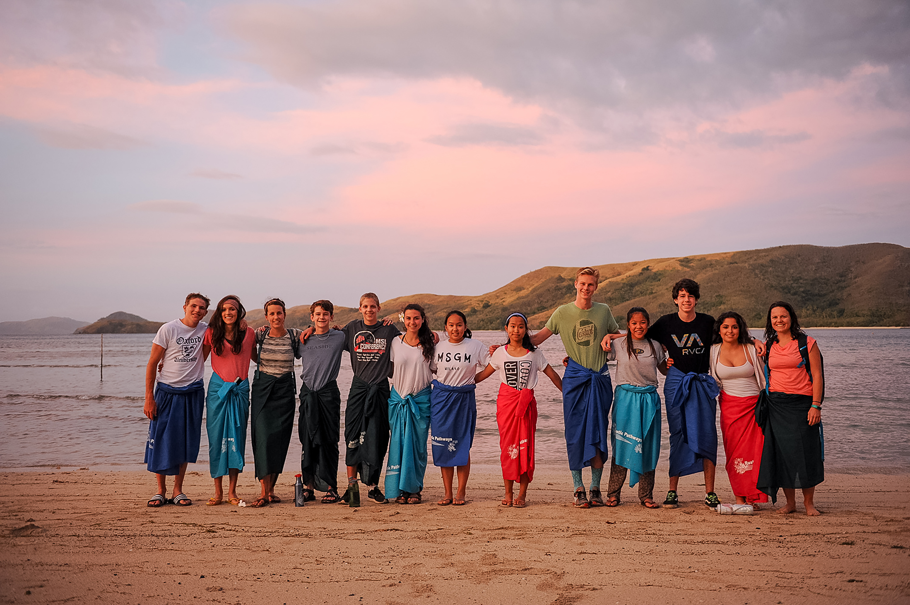 Students in traditional wraps pose together on a beach at sunset