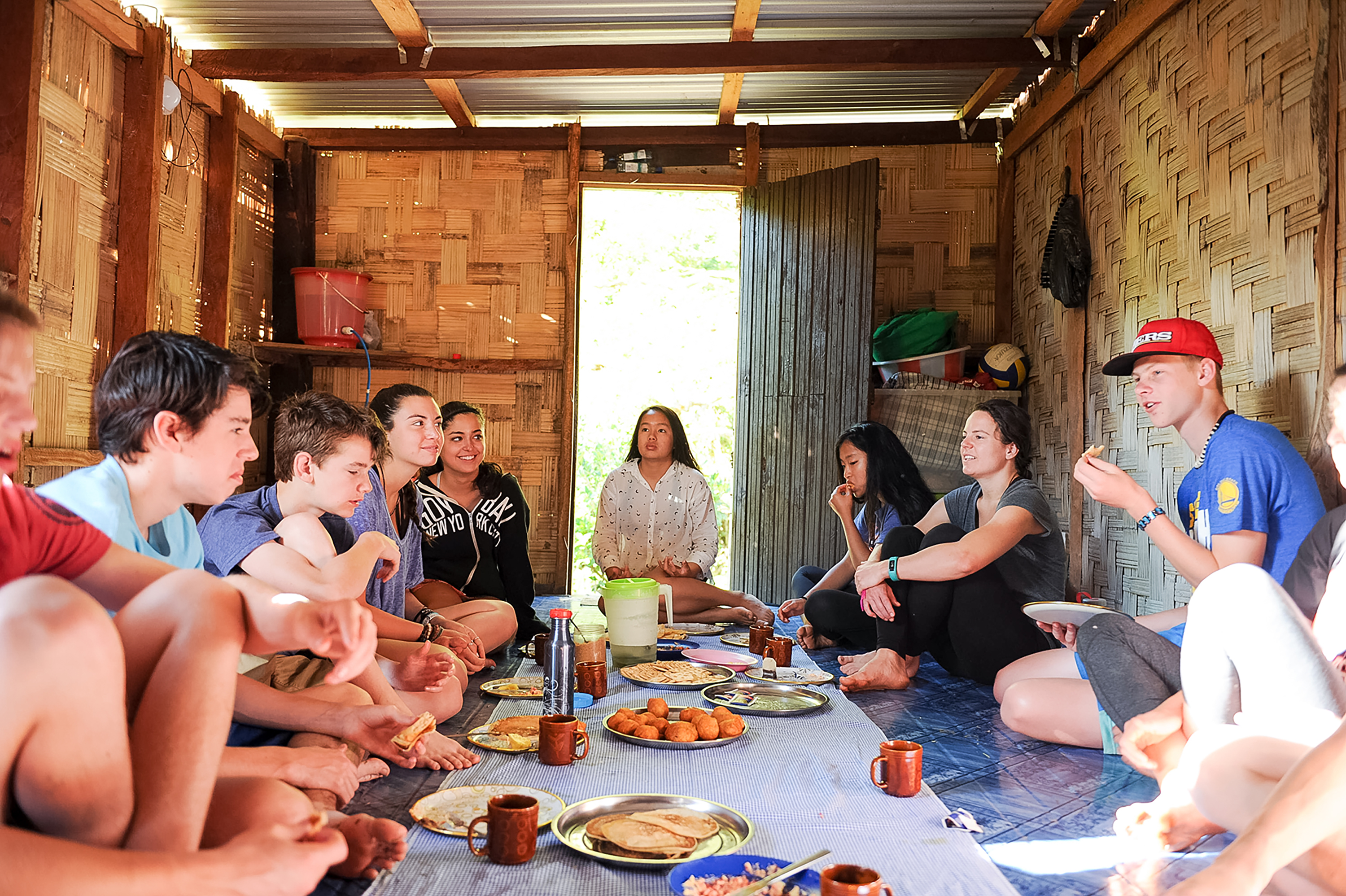 Students share a meal while seated on the floor of a bamboo hut with a local host