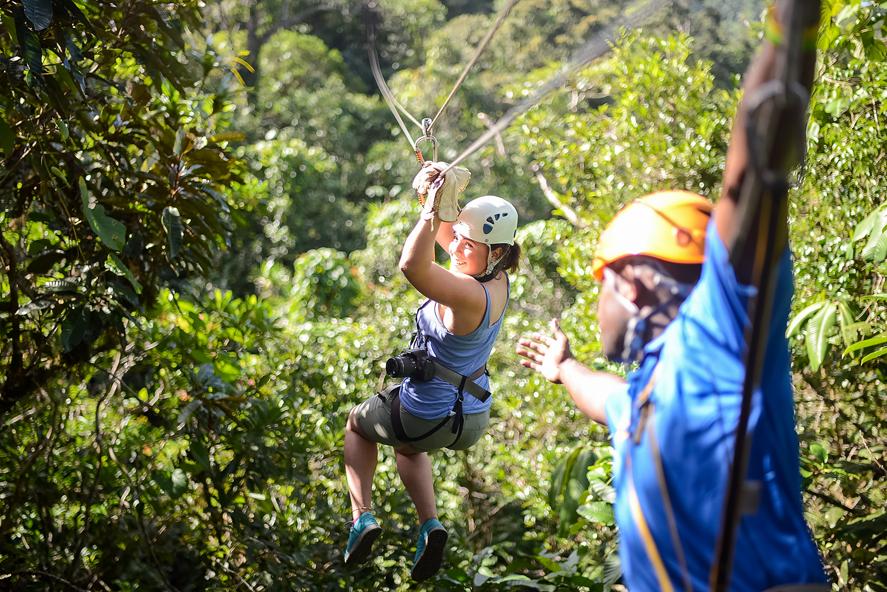 A student ziplines through the forest canopy wearing a helmet and harness