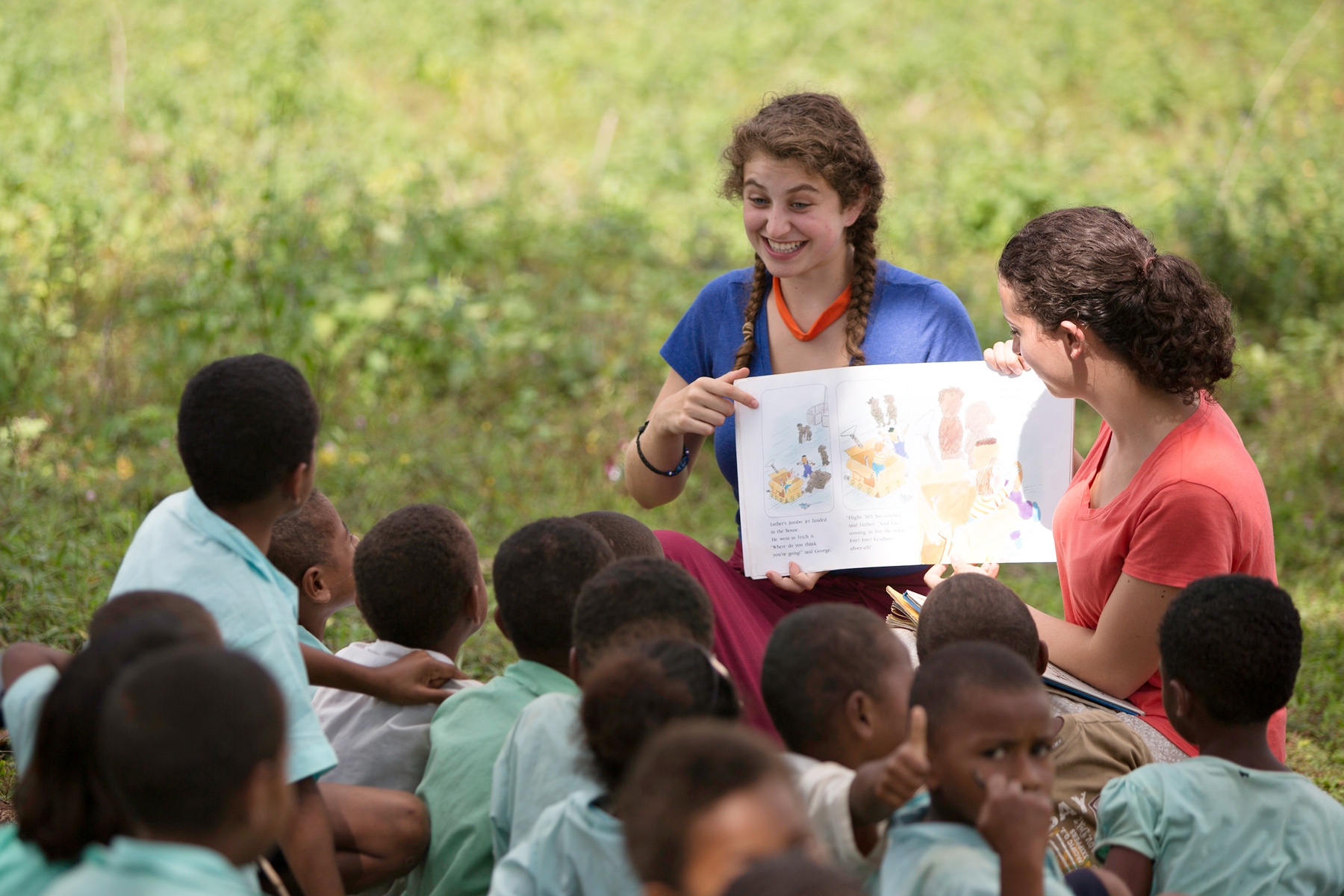Students read a picture book aloud to local children seated in a grassy field